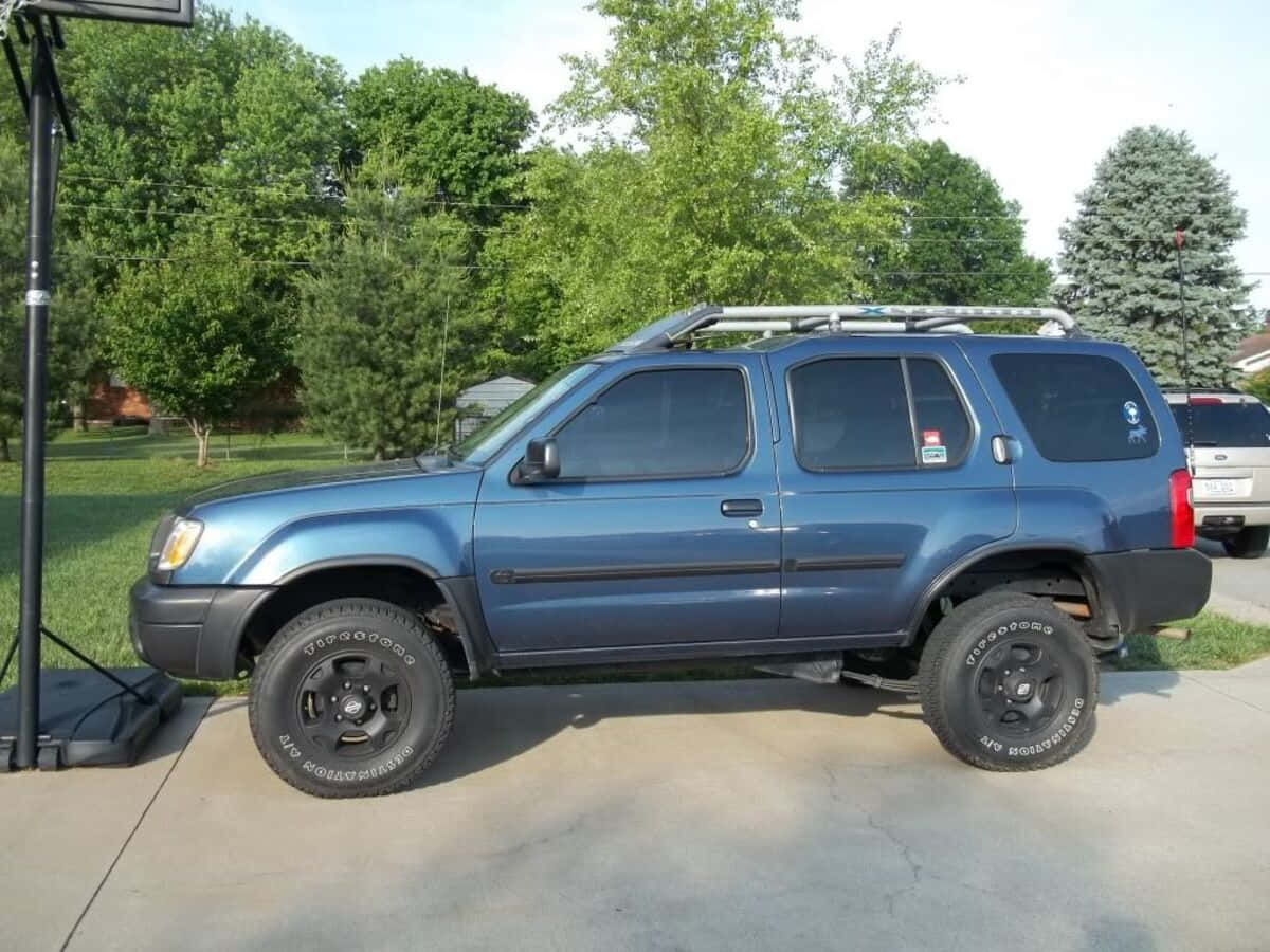 Nissan Xterra 桌布 壁紙 — 一輛升高底盤的藍色SUV，配備黑色輪圈和車頂行李架，停放在混凝土車道上，周圍環繞著綠樹。