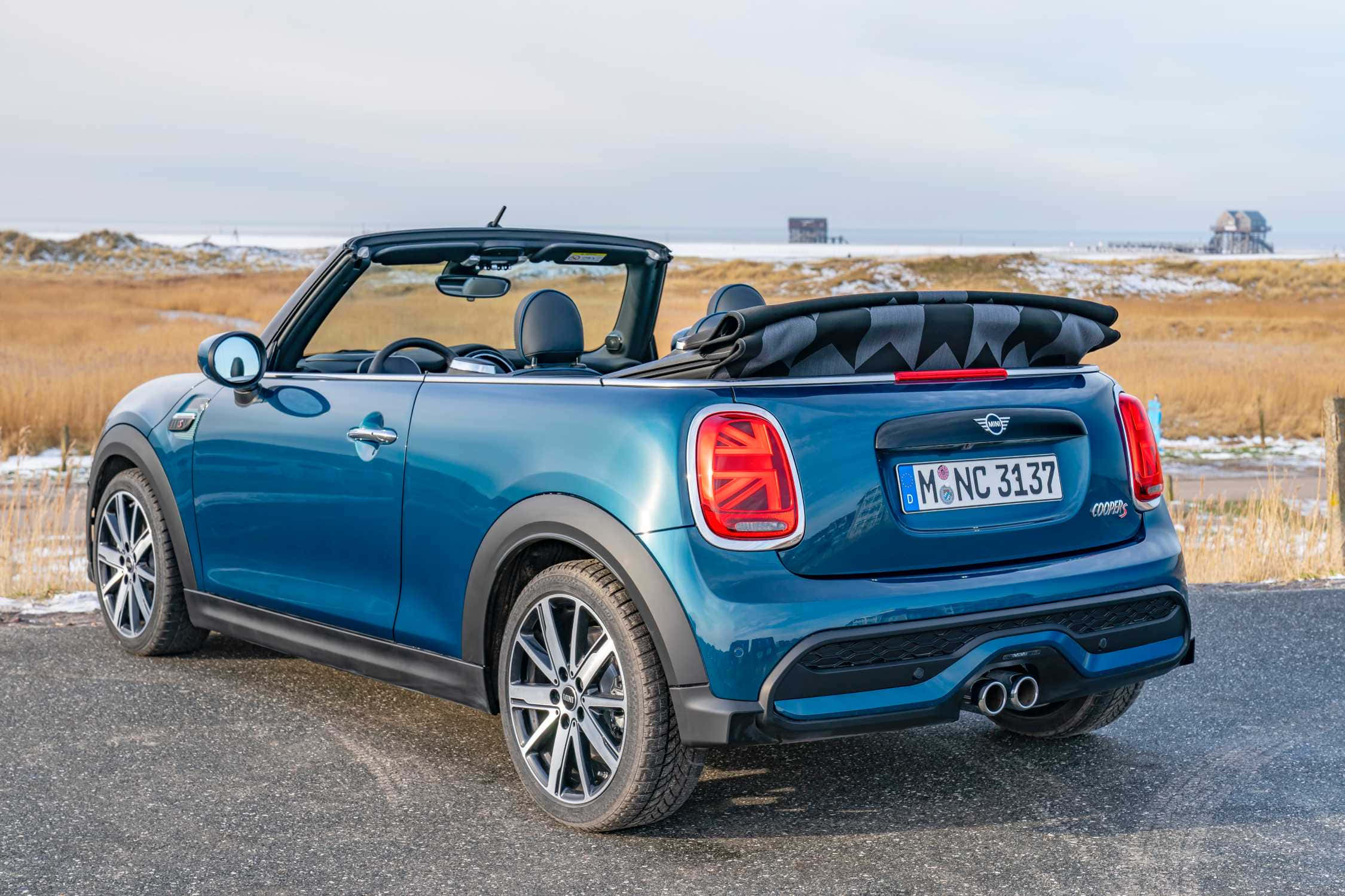 Mini Cooper S Convertible 桌布 壁紙 — 一輛鮮藍色的MINI Cooper Convertible停泊在碎石路上，周圍是雪地田野和遠處的建築物，天空多雲。