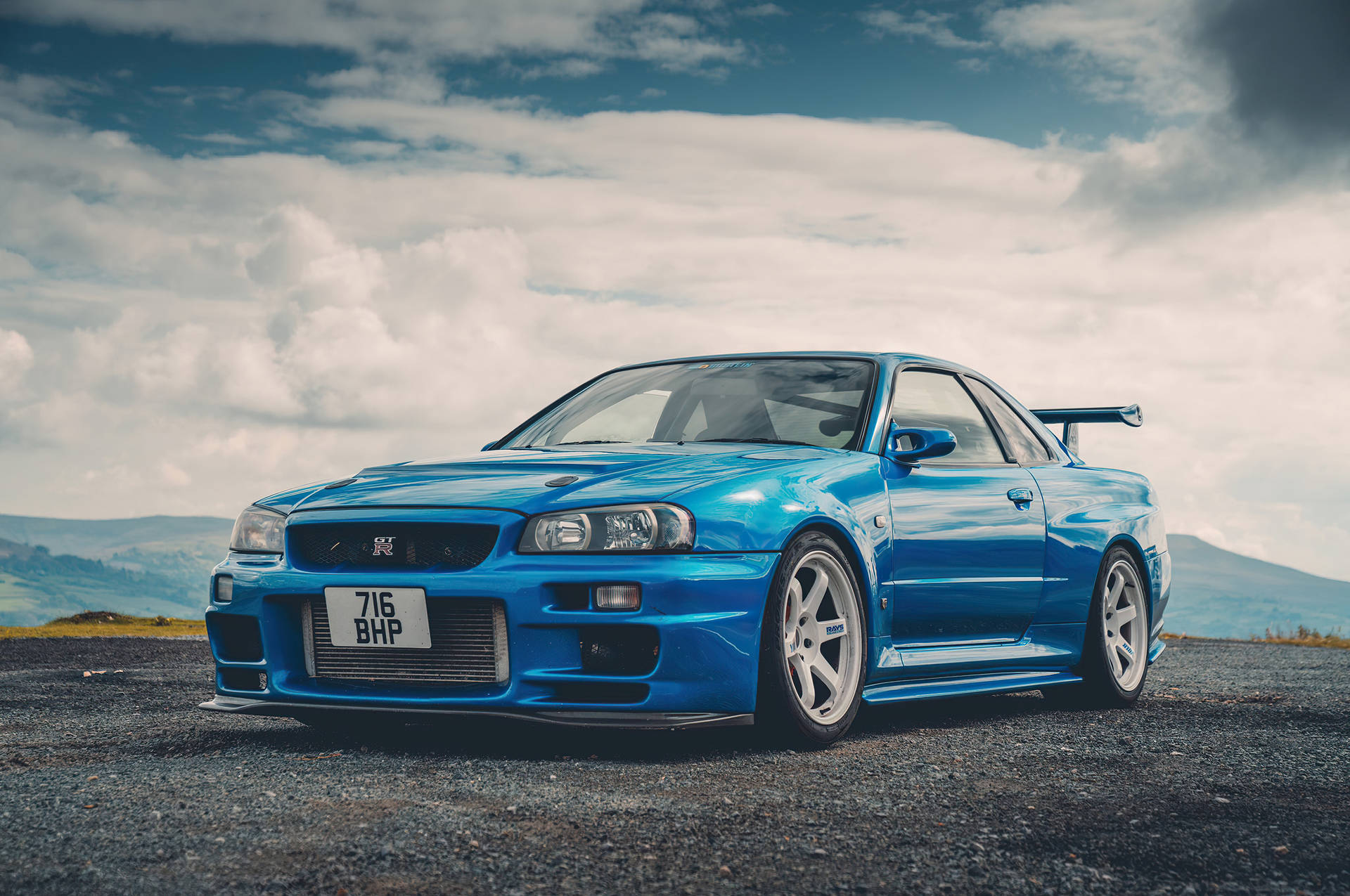藍色日產Skyline GT-R在碎石路上，背景有丘陵和多雲天空。 — 天際線 壁紙
