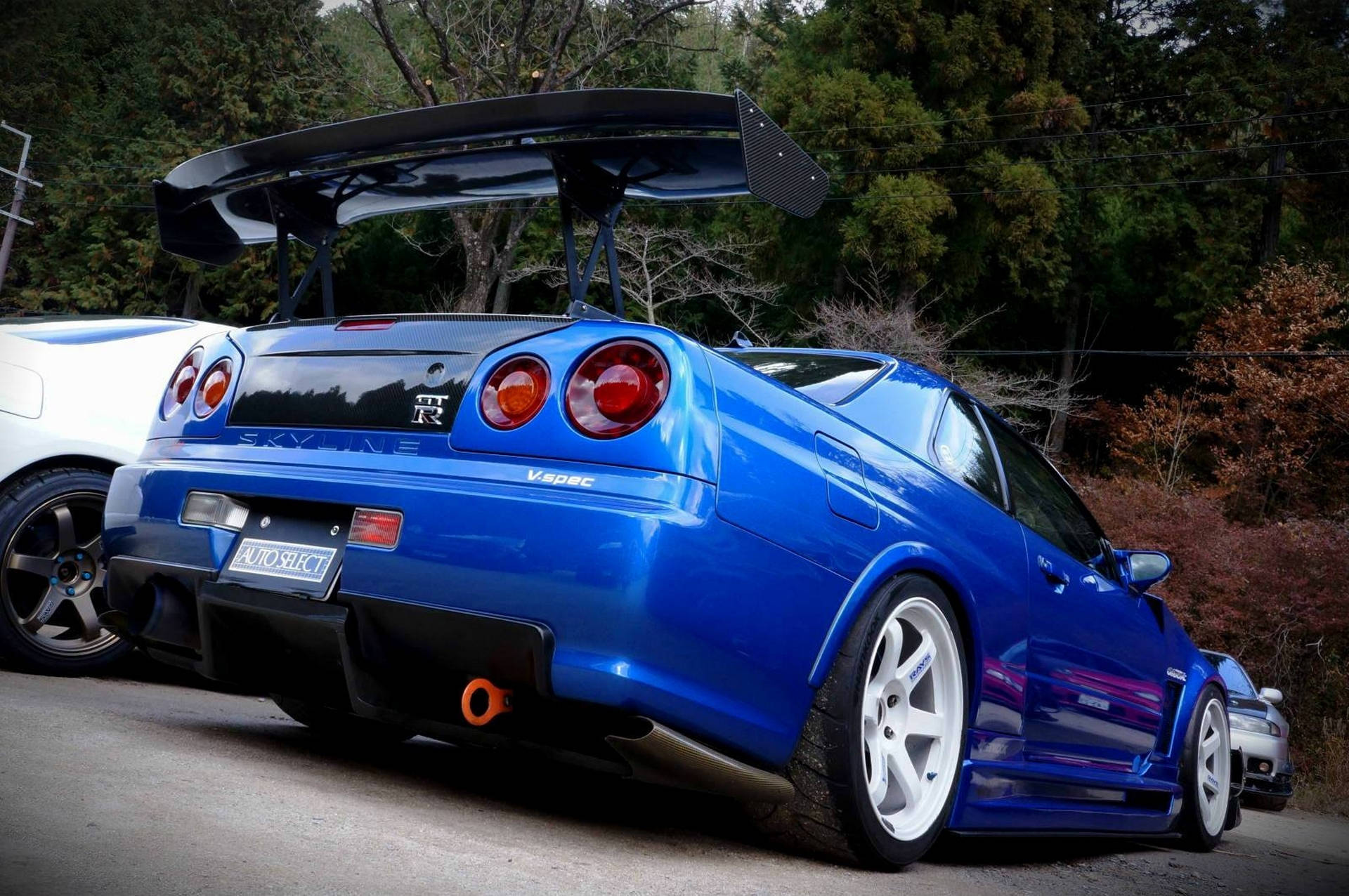 藍色日產Skyline R34 V-Spec，帶有大型後尾翼，停放在秋樹附近的道路上。 — 日產 Skyline GTR R34 壁紙