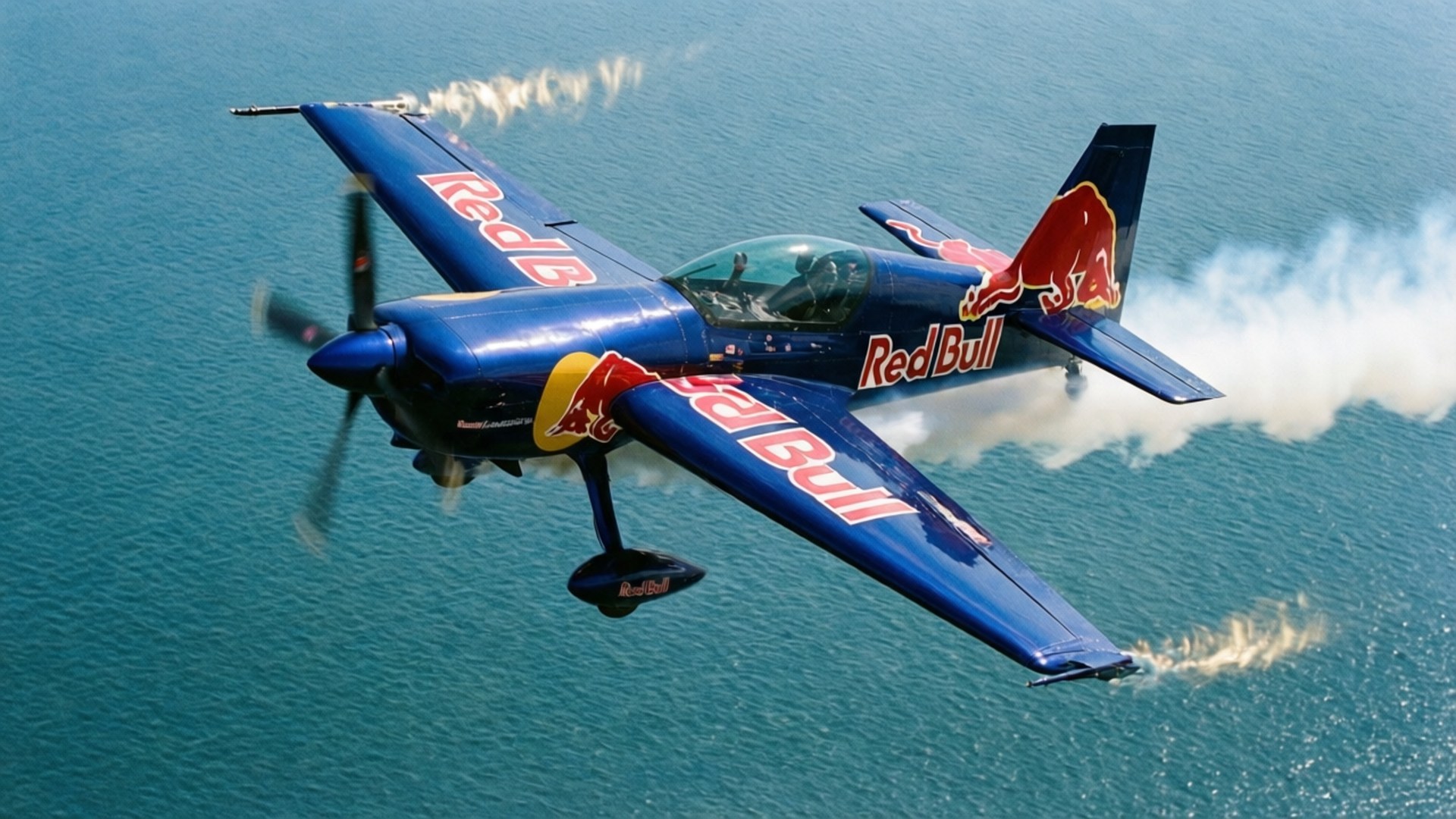 Air Race Over Water 壁紙 — 一架帶有紅色標誌的藍色Red Bull特技飛機在藍綠色海水上空急轉彎，留下白色煙霧痕跡。
