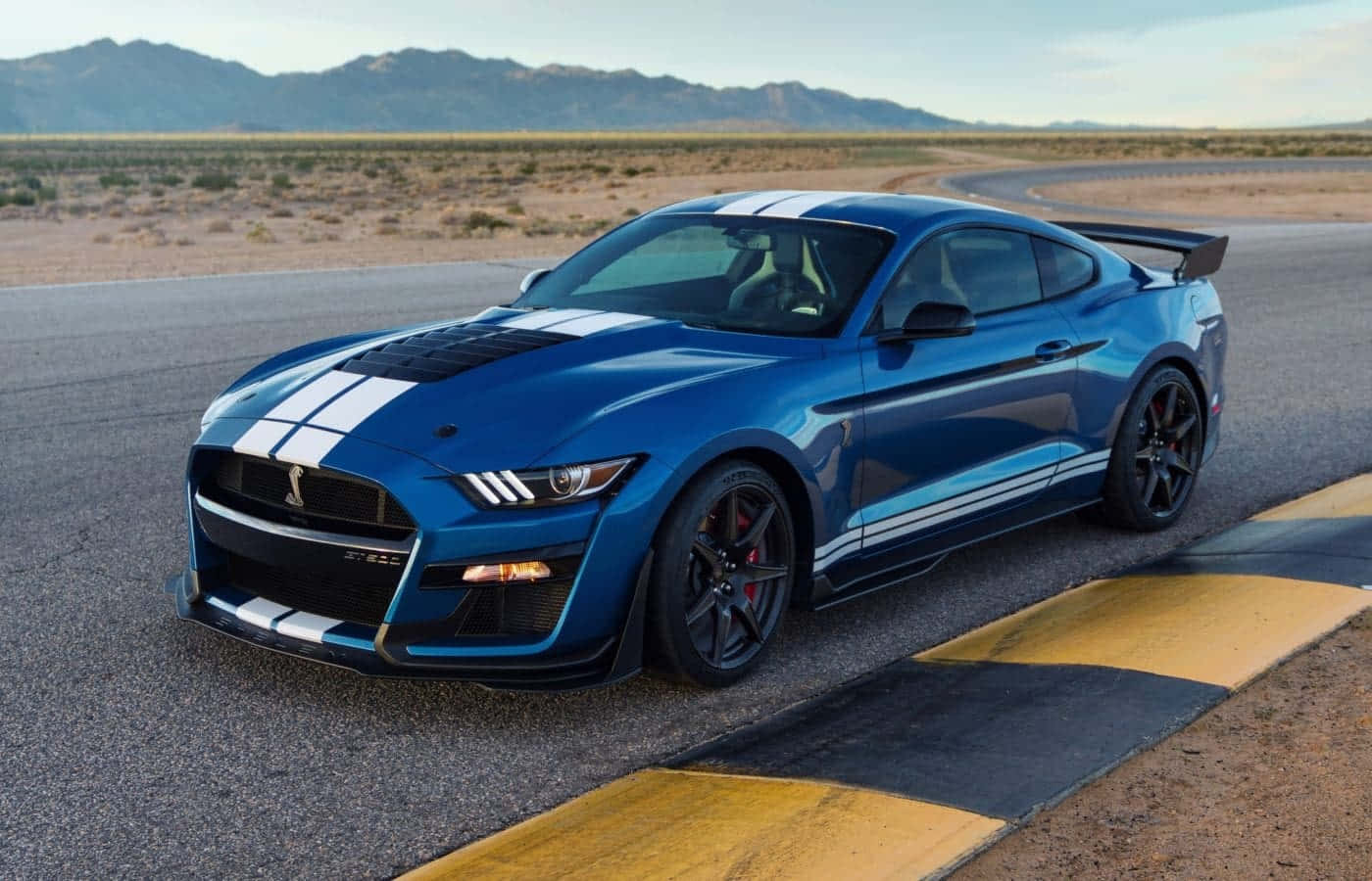 壁紙 — 一輛充滿活力的藍色Shelby GT500跑車，帶有白色賽車條紋，在晴朗的天空下沿著沙漠賽道飛馳。
