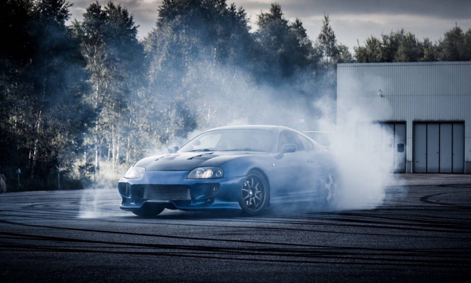 藍色跑車在倉庫附近的瀝青場地上在煙霧中飄移。 — Toyota Supra 壁紙
