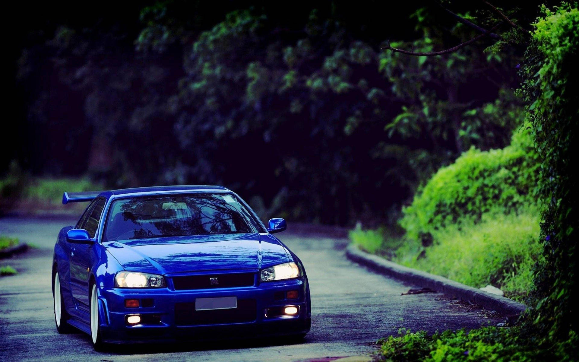 藍色跑車在黃昏的森林道路上，車頭燈亮著。 — 日產Skyline GTR R33壁紙 壁紙