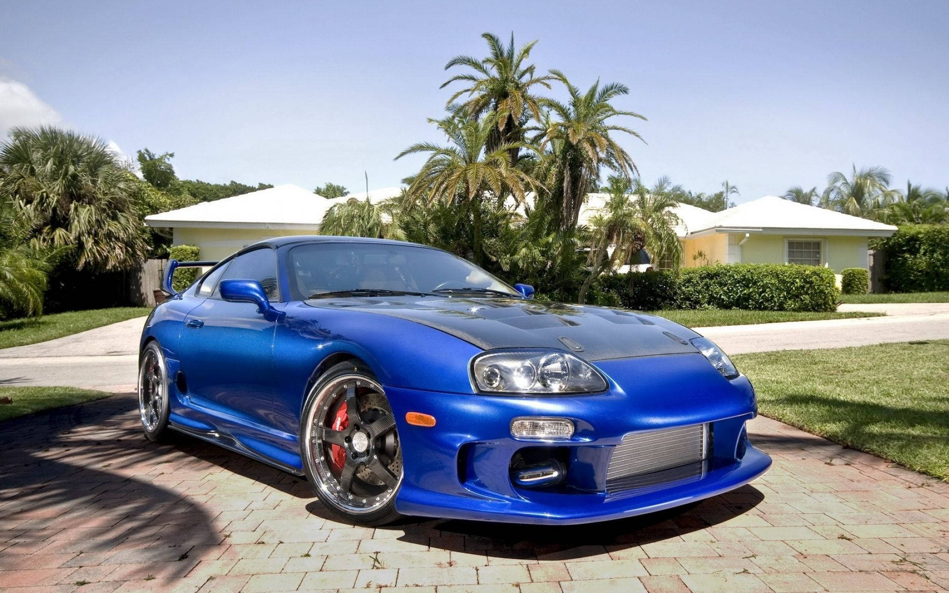 藍色豐田Supra停放在磚砌車道上，背景有棕櫚樹和房屋。 — Toyota Supra 壁紙