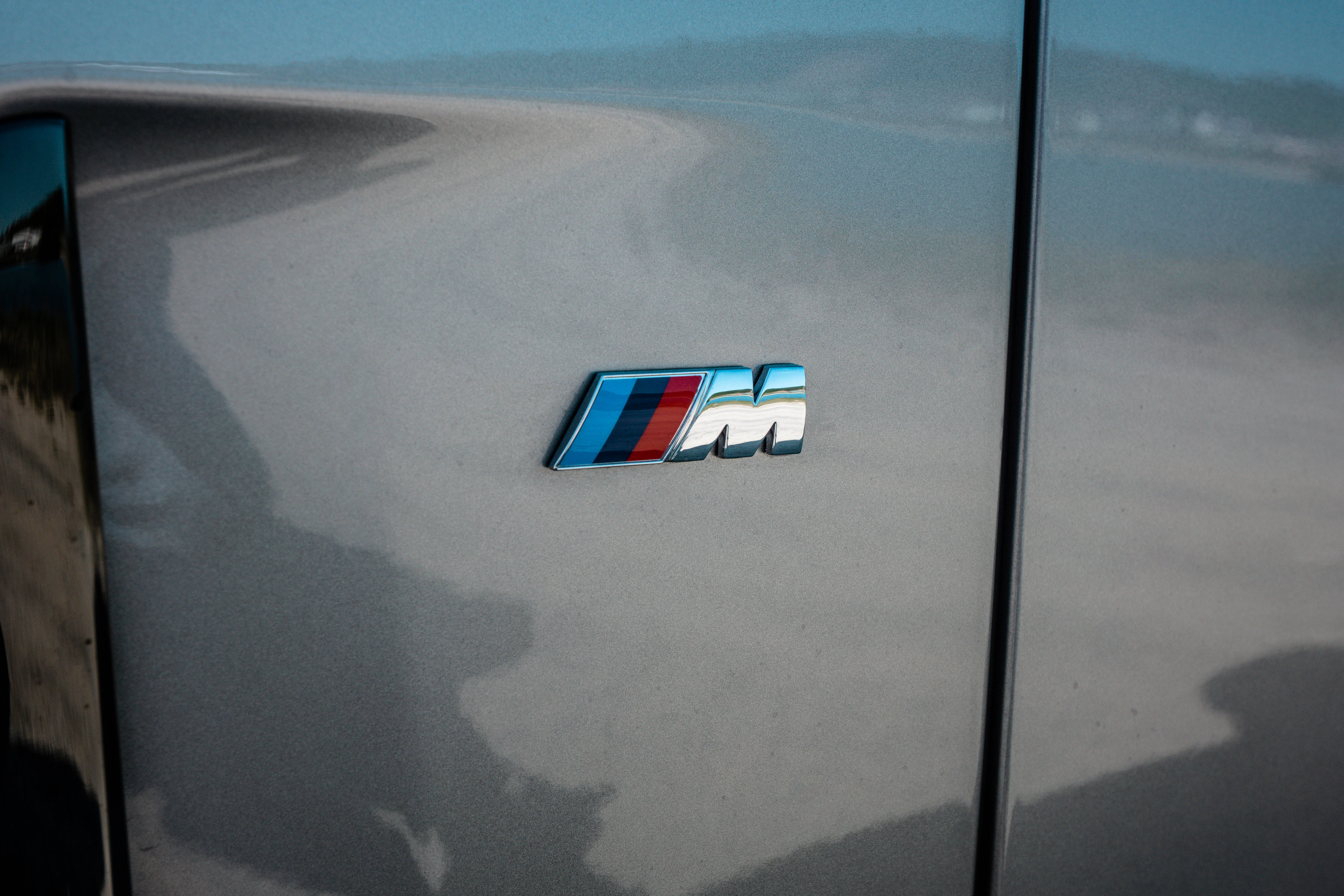 m 桌布 壁紙 — BMW M 徽章特寫，位於時尚車門上，反射出海岸道路和遠山，在柔和日光下。