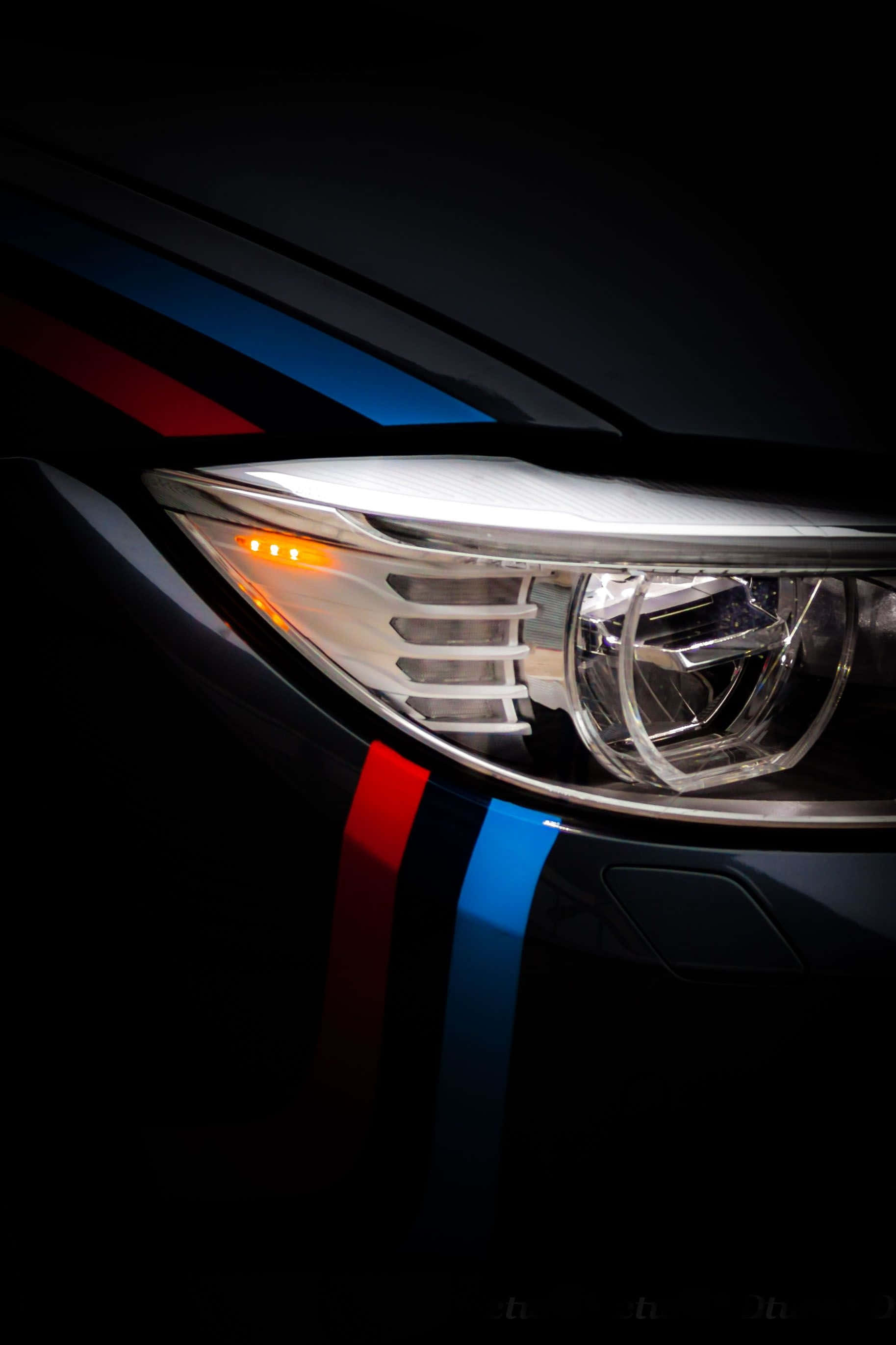 Bmw Android 桌布 壁紙 — BMW M 車頭燈的特寫，在黑色背景上發出光芒，帶有紅色、藍色和白色的賽車條紋。