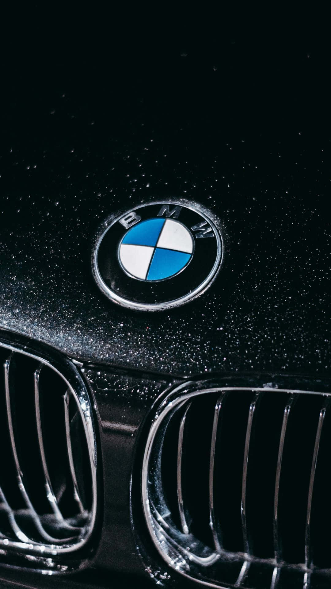 Android Bmw 桌布 壁紙 — BMW 標誌在濕黑色引擎蓋上閃耀的特寫，帶有鍍鉻格柵細節。