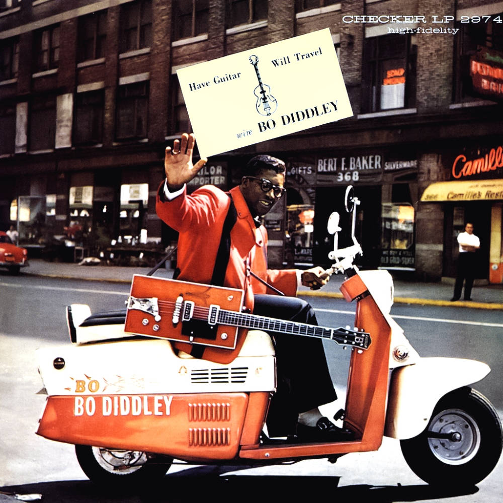 吉他動圖 桌布 壁紙 — Bo Diddley 微笑著騎著橙色滑板車，背著吉他，手持寫有「Have Guitar Will Travel」的牌子。