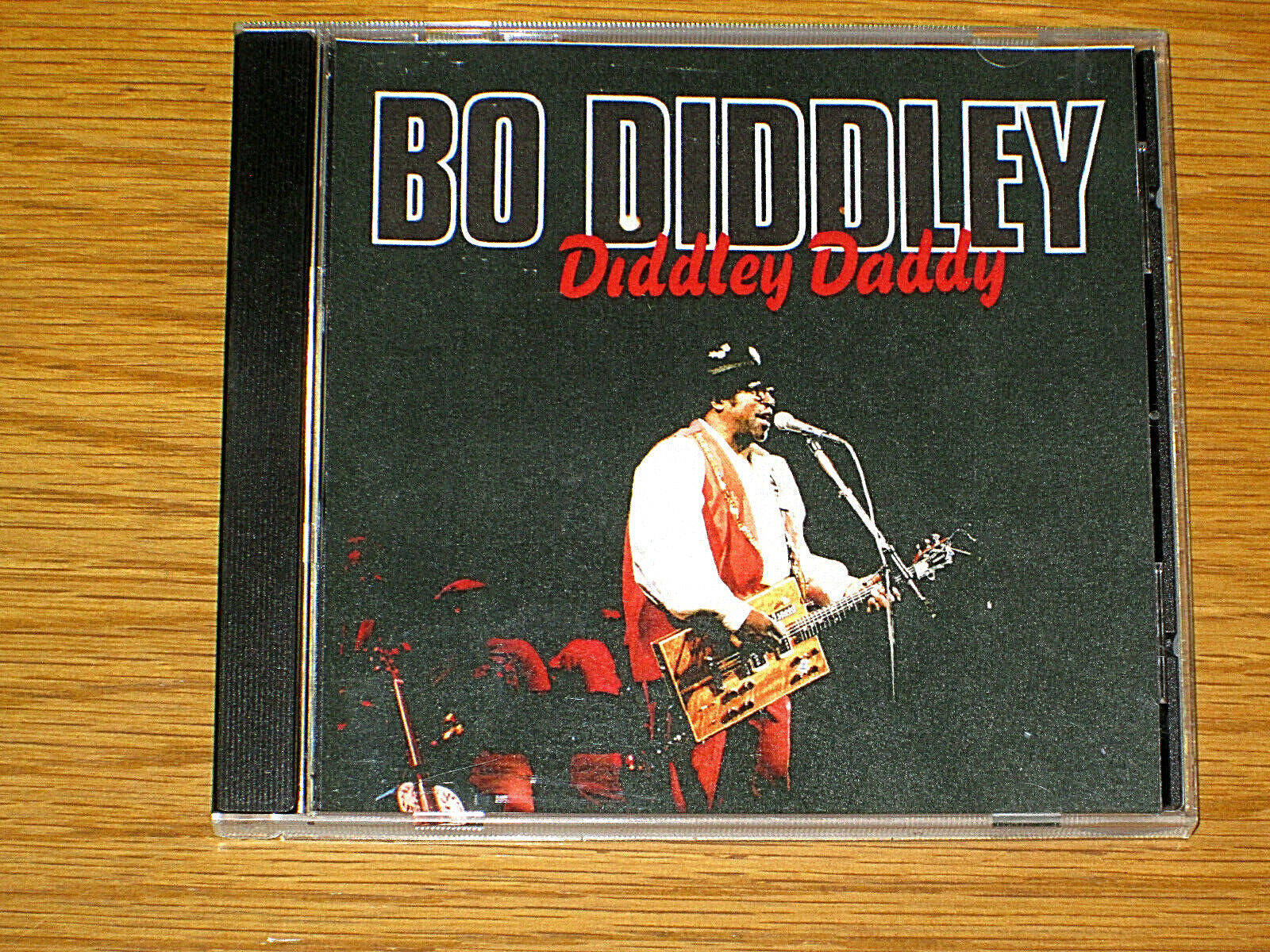 Album Cover 桌布 壁紙 — Bo Diddley 的《Diddley Daddy》CD 盒放在木質表面上，顯示藝術家正在彈吉他並對著麥克風唱歌。
