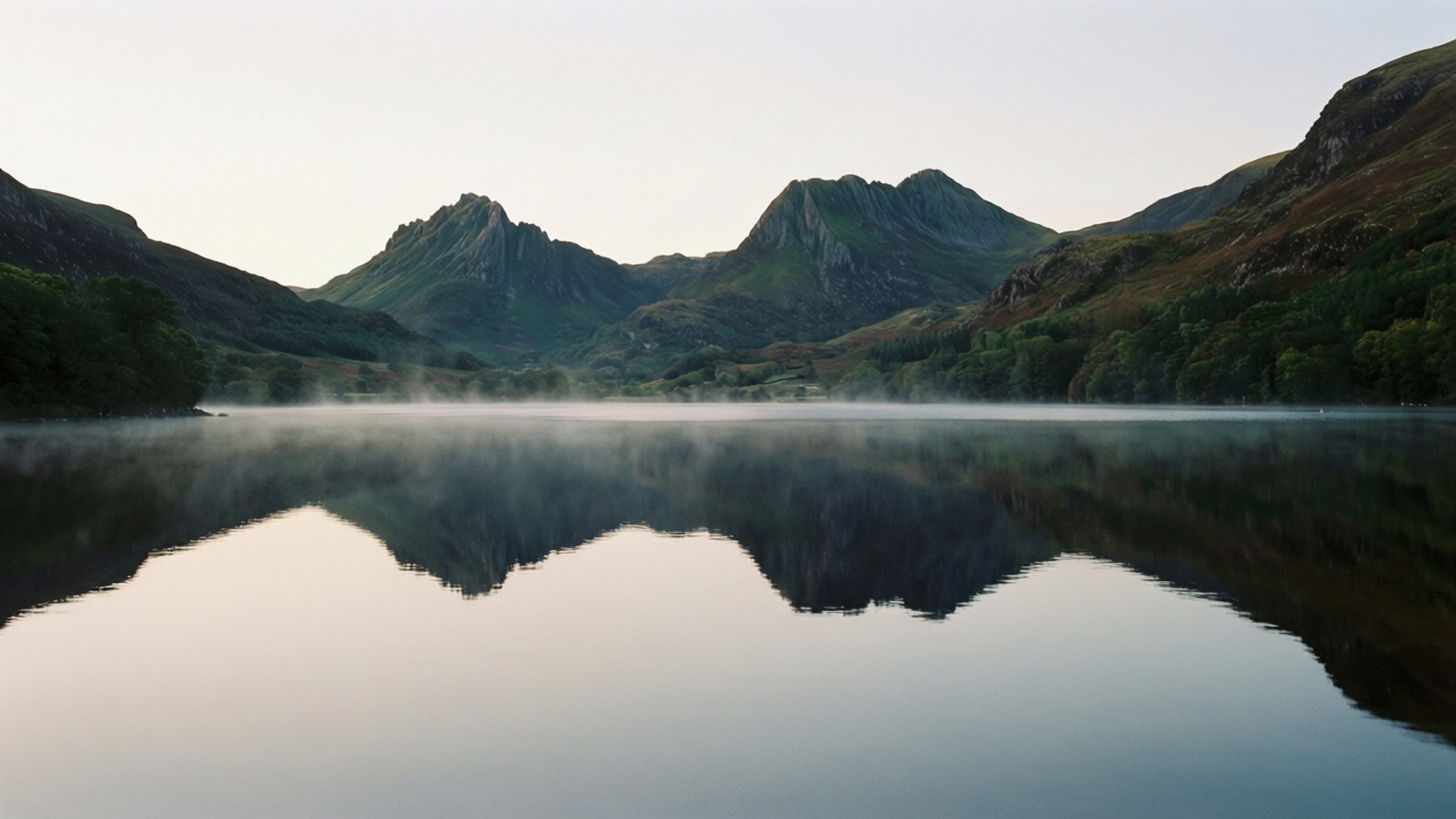 Connemara Lake Mist 壁紙 — 薄霧黎明，平靜的湖面映照著參差不齊的綠色山脈，天空柔和蒼白。