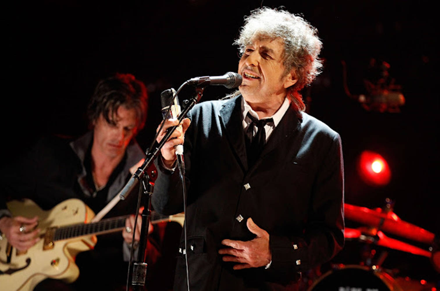 黑白 桌布 壁紙 — Bob Dylan 在舞台上熱情地對著麥克風唱歌，身後有吉他手在紅色舞台燈光下演奏。