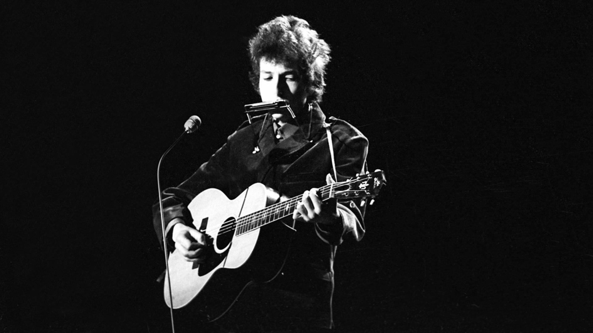 黑白 桌布 壁紙 — Bob Dylan 在舞台上表演，手持木吉他和口琴，在黑色背景的戲劇性聚光燈下。