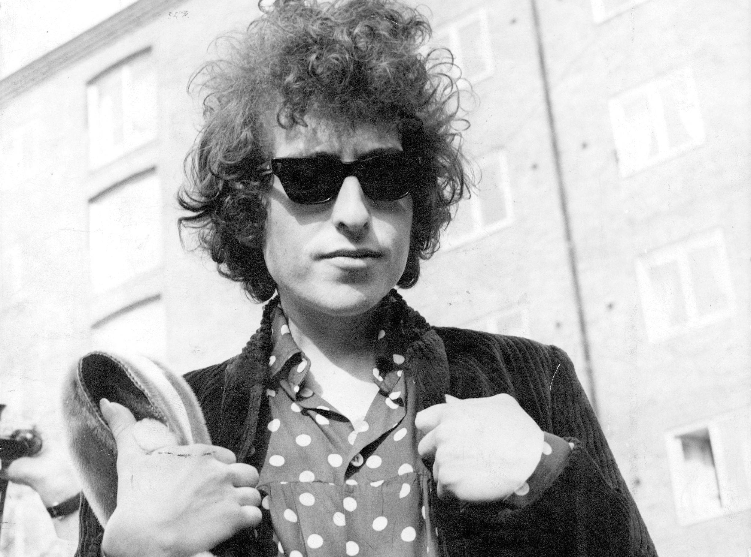 黑白 桌布 壁紙 — Bob Dylan 自信地以黑白風格站立，戴著太陽眼鏡，穿著波點襯衫，手持帽子，背景為建築物。