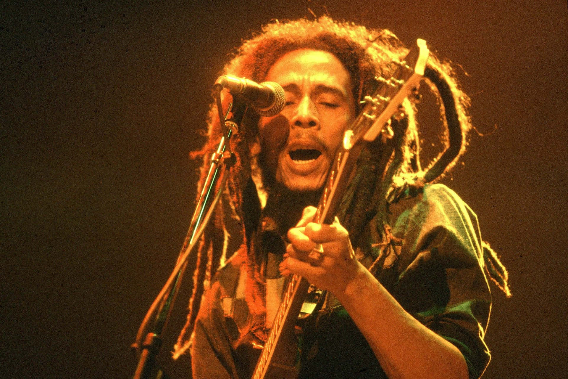 Bob Marley 桌布 壁紙 — Bob Marley 在溫暖的舞台燈光下，熱情地對著麥克風唱歌，同時彈奏吉他。