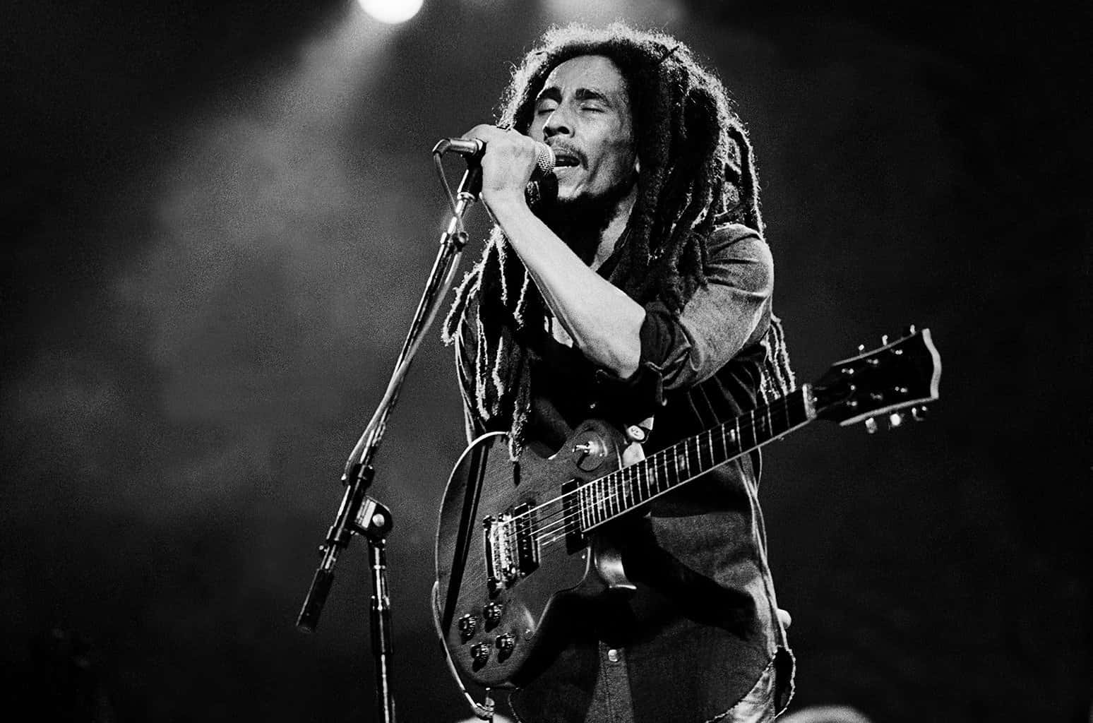 Bob Marley 桌布 壁紙 — Bob Marley 在黑白舞台燈光下，熱情地對著麥克風唱歌，同時手持吉他。