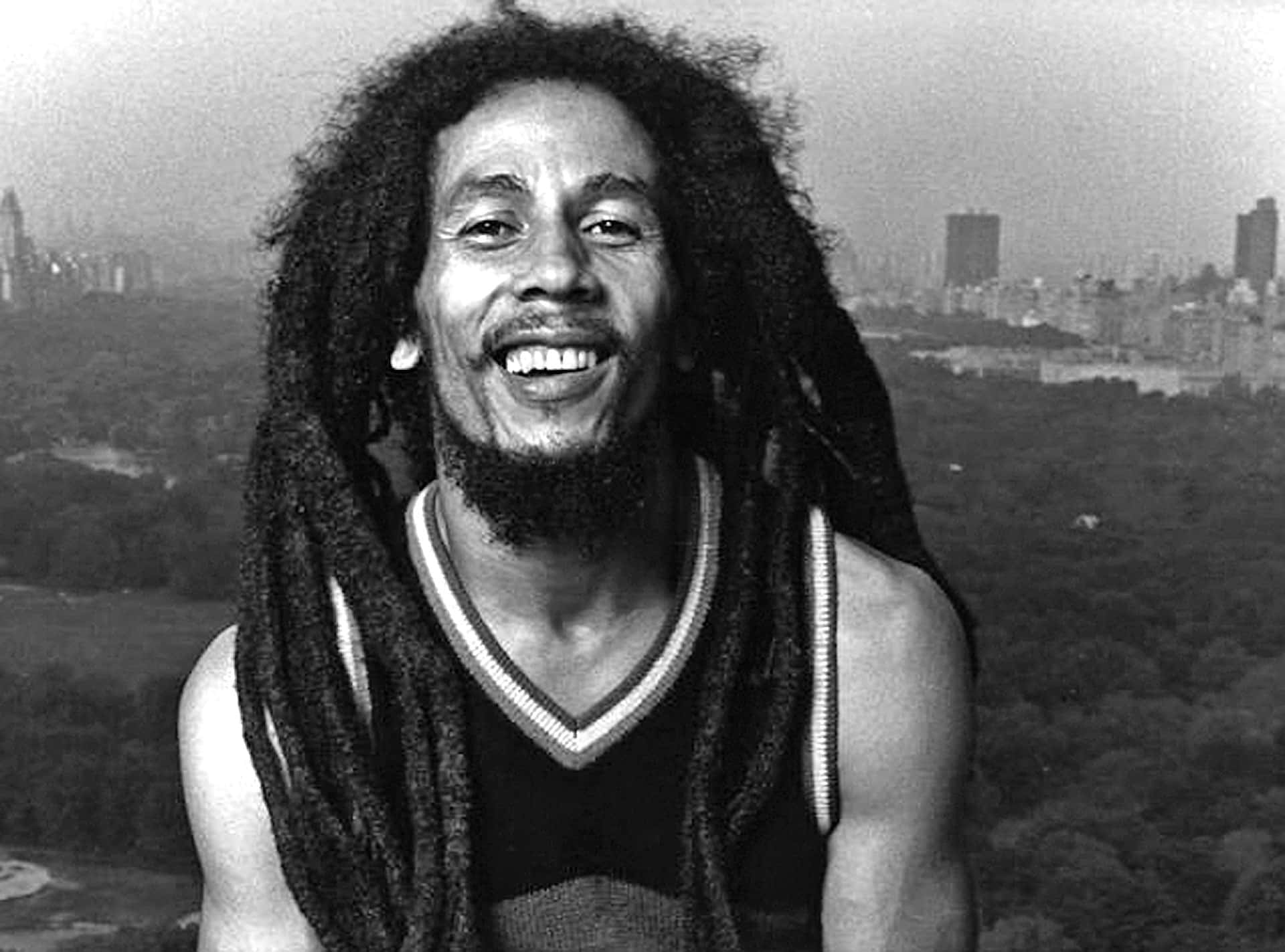 Bob Marley 桌布 壁紙 — Bob Marley 溫暖地微笑，長髮綹框住他的臉龐，背景是朦朧的紐約天際線和下方的綠色公園。
