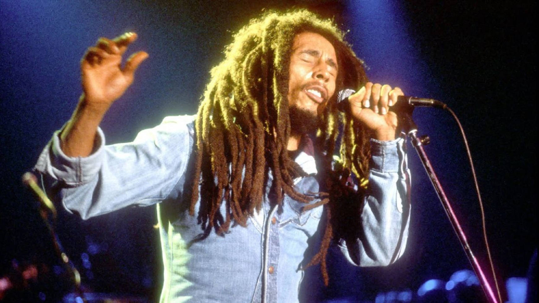 音樂 桌布 壁紙 — Bob Marley 閉上眼睛，高舉雙臂，在充滿活力的舞台燈光下熱情地對著麥克風唱歌。