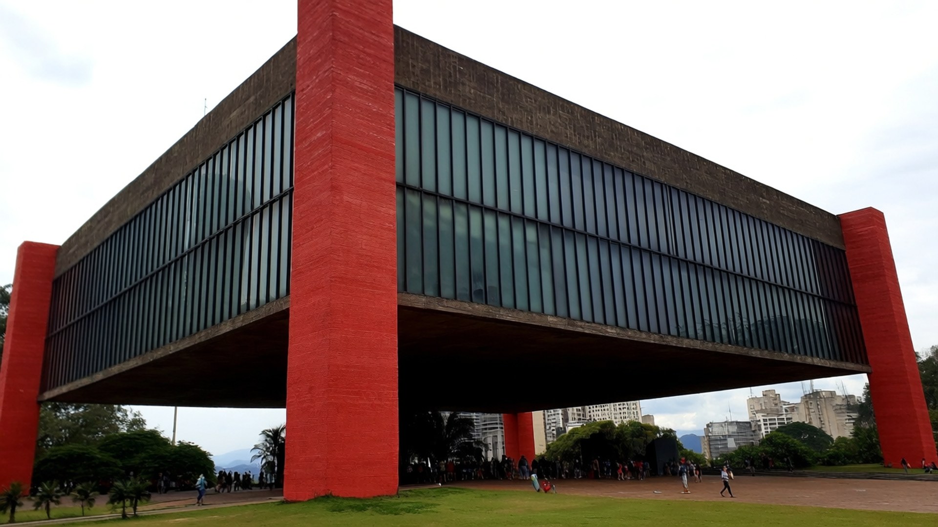 MASP Modern Icon 壁紙 — 一座大膽的現代主義建築，擁有巨大的紅色柱子和深色玻璃外牆，聳立在陰天下的綠色草坪上。