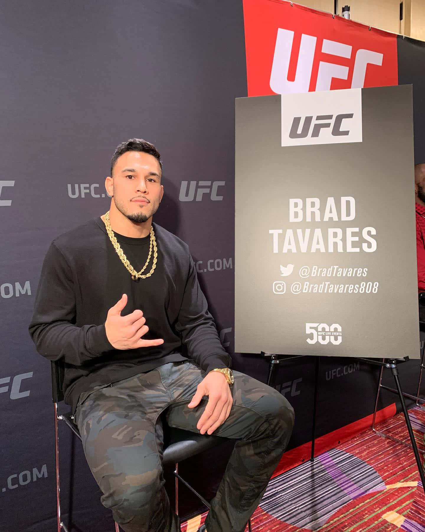 UFC 桌布 壁紙 — Brad Tavares 自信地坐在 UFC 活動的椅子上，身穿黑色襯衫和迷彩褲，戴著金鏈。