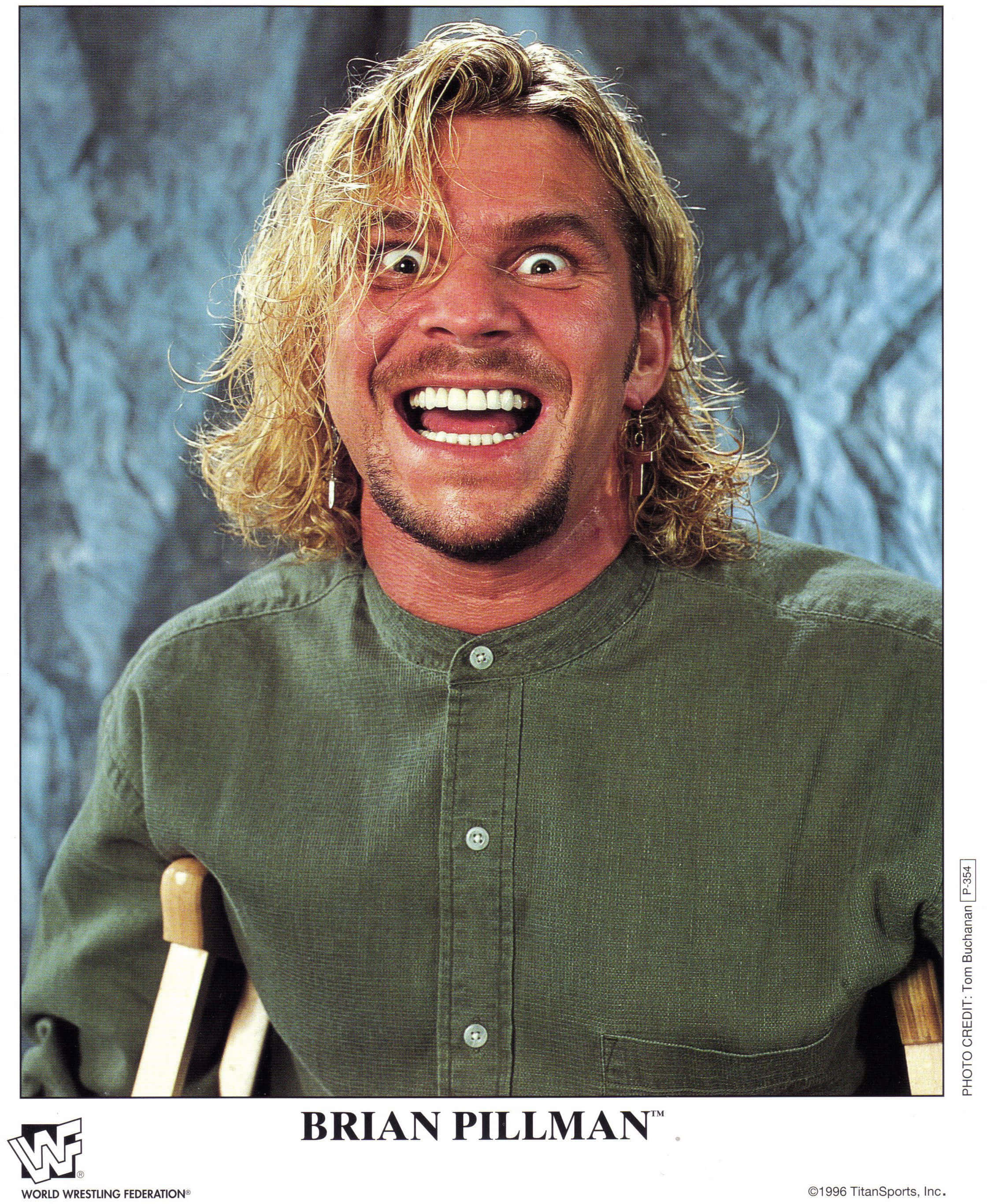 WWE 桌布 壁紙 — Brian Pillman 在有紋理的藍色背景前，瞪大雙眼、一頭凌亂金髮，咧嘴狂笑。