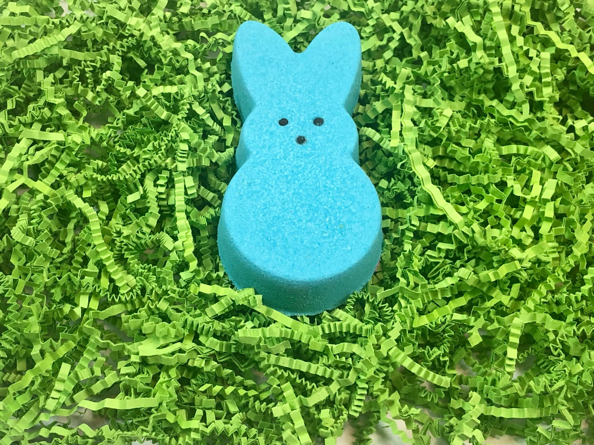  壁紙 — 一個亮藍色兔子形狀的浴球放在充滿活力的綠色碎紙中。