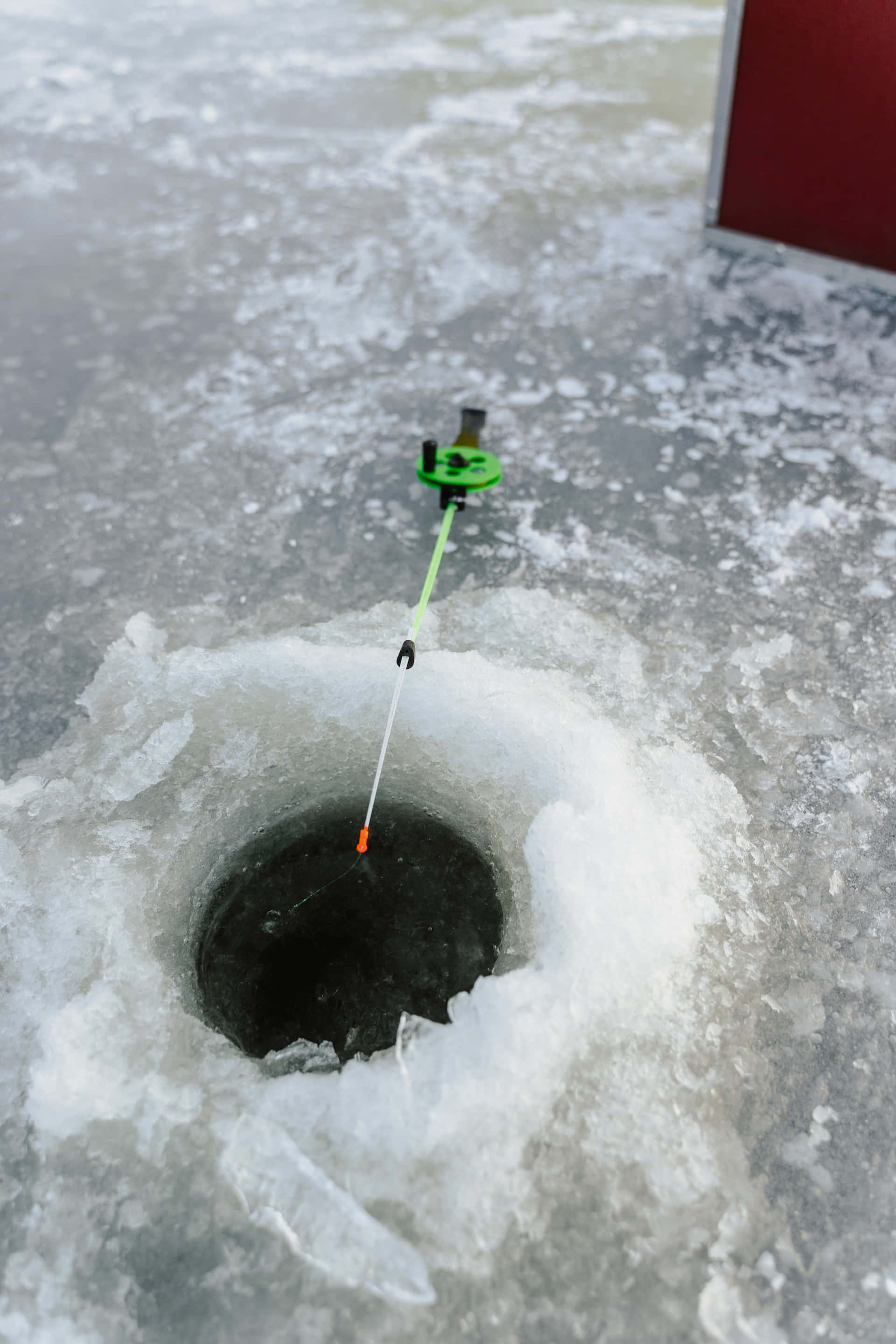 Ice Fishing 桌布 壁紙 — 一個亮綠色的冰釣提示器在陰天之下，立於一個紋理冰凍湖面的暗洞之上。
