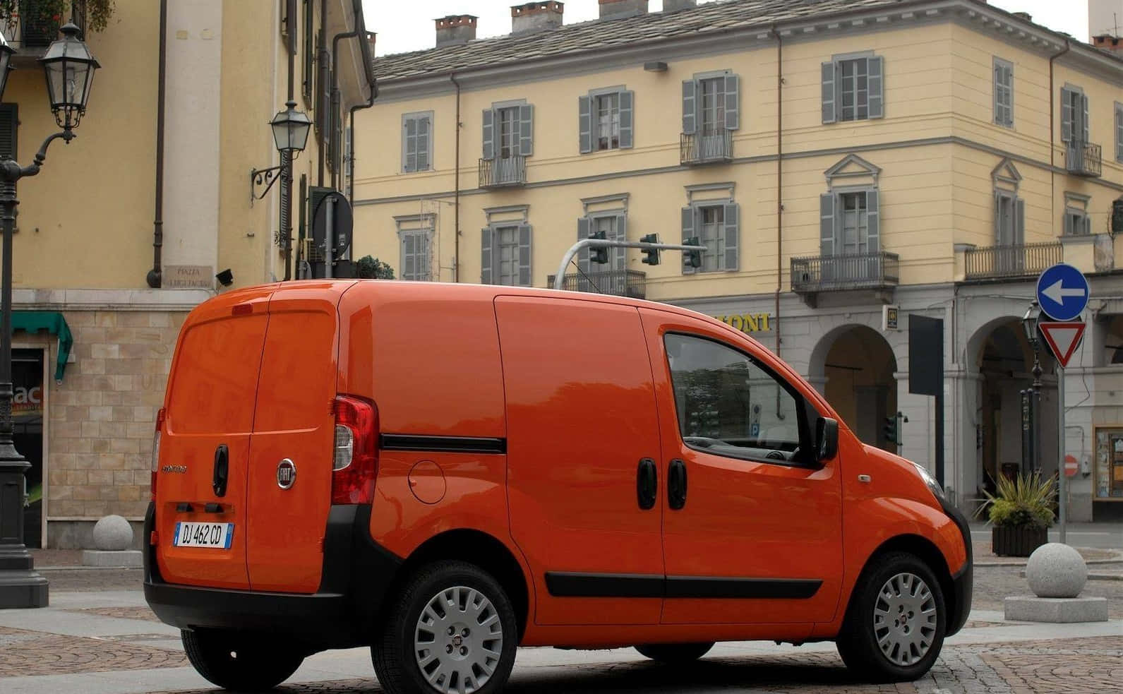 Fiat Fiorino 桌布 壁紙 — 一輛亮橙色的菲亞特貨車停泊在鵝卵石街道上，前面是帶有灰色百葉窗的歷史黃色建築。