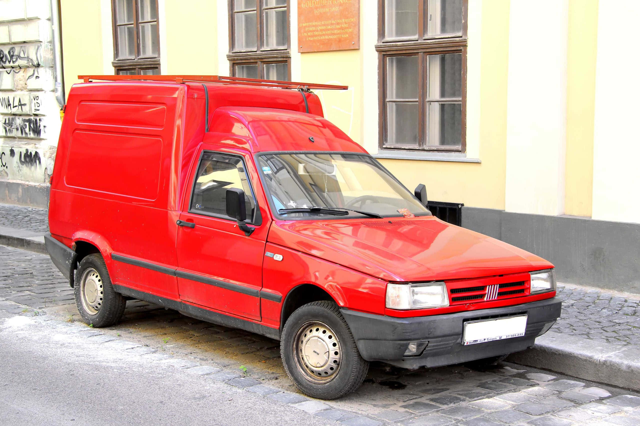 Fiat Fiorino 桌布 壁紙 — 一輛鮮紅色復古飛雅特貨車停泊在鵝卵石街道上，旁邊是一棟帶有塗鴉的黃色建築。