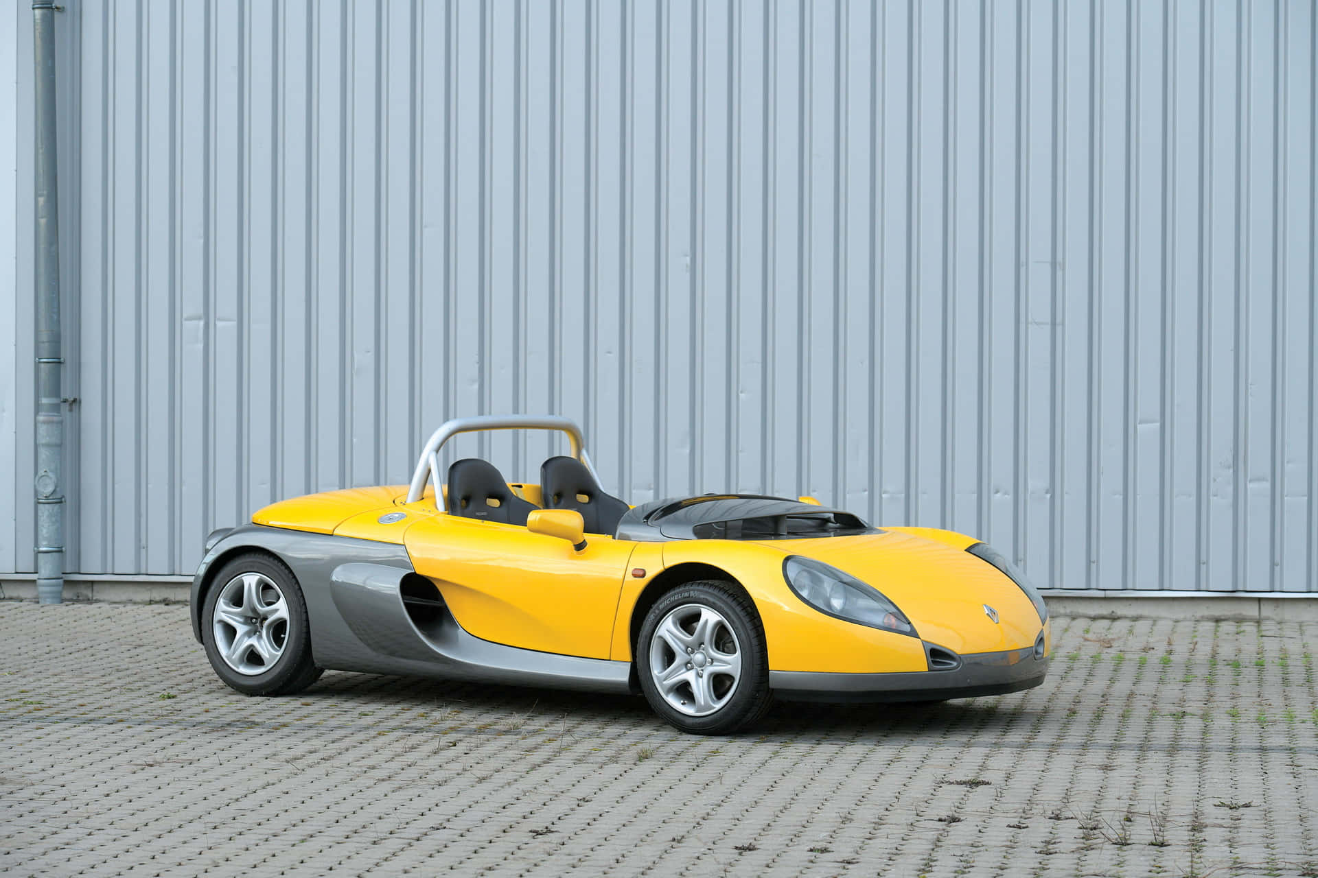 Renault Sport Spider 桌布 壁紙 — 一輛亮黃色和銀色的雙座跑車停放在鋪砌路面上，背景是波紋金屬牆。
