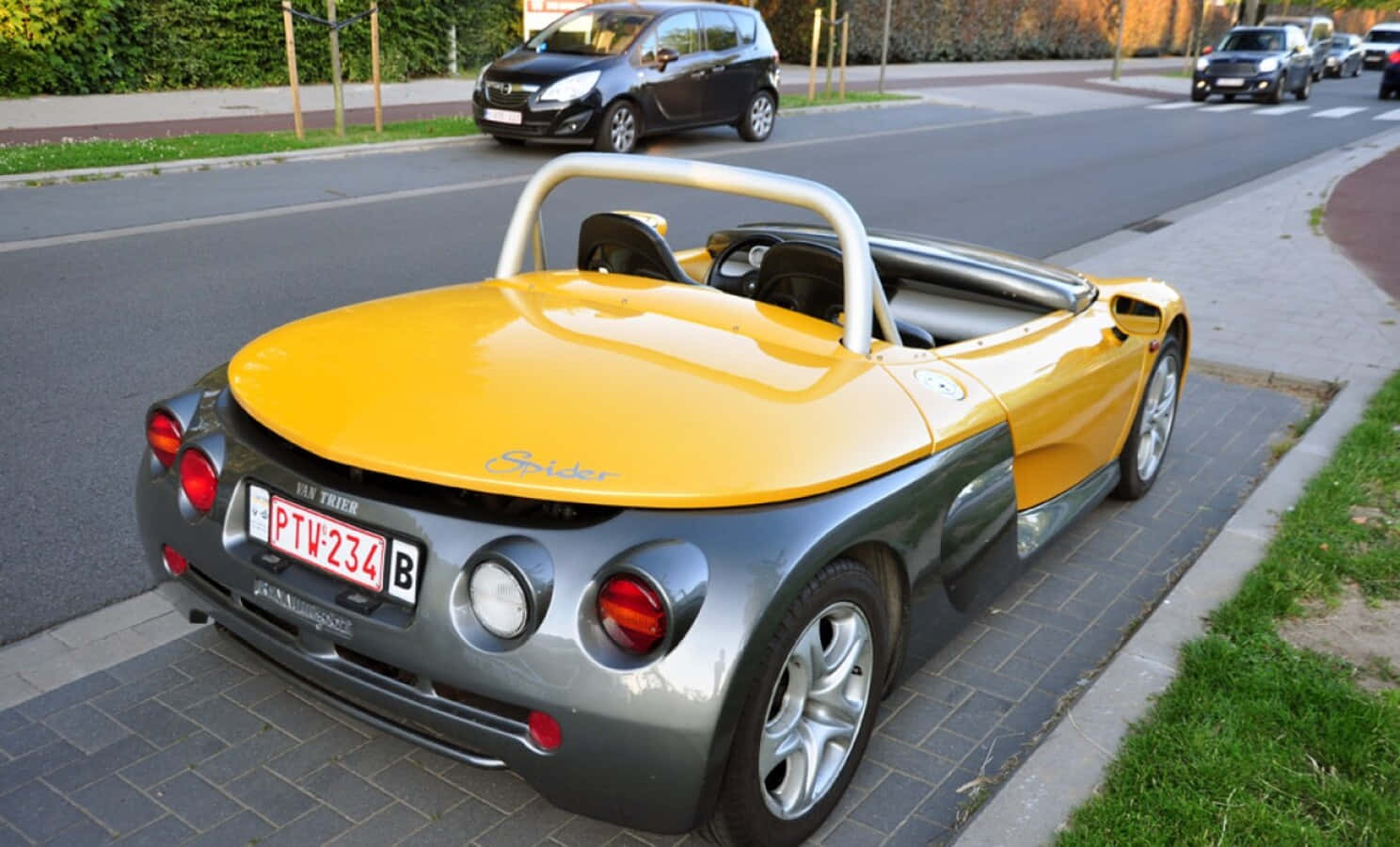 Renault Sport Spider 桌布 壁紙 — 一輛亮黃色和銀色的TVR Spider敞篷車停放在磚砌人行道上，旁邊是一條有其他車輛和綠化的街道。