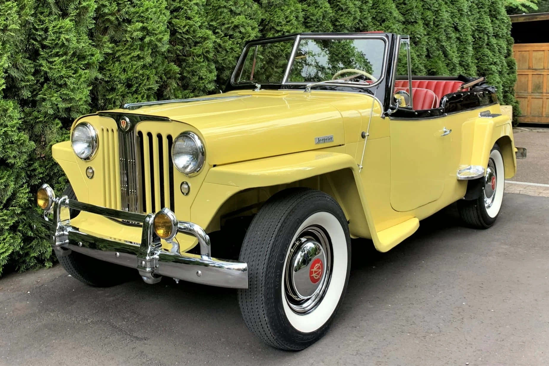 Jeepster 桌布 壁紙 — 一輛亮黃色復古敞篷車，紅色內裝，停在鋪砌車道旁茂密的綠色樹籬邊。