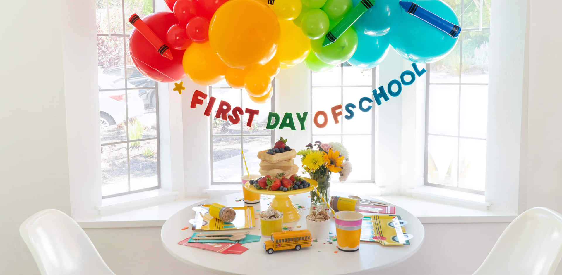 壁紙 — 一張明亮裝飾的桌子，帶有彩虹氣球拱門和「FIRST DAY OF SCHOOL」橫幅，放在陽光充足的窗邊。