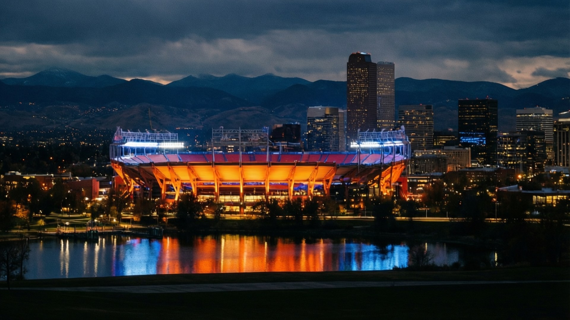 Denver Night Skyline 壁紙 — 一個明亮照明的體育場在黃昏時發出橙色和藍色的光芒，其倒影在水面上閃爍，前方是城市天際線和遠處的山脈，天空多雲。