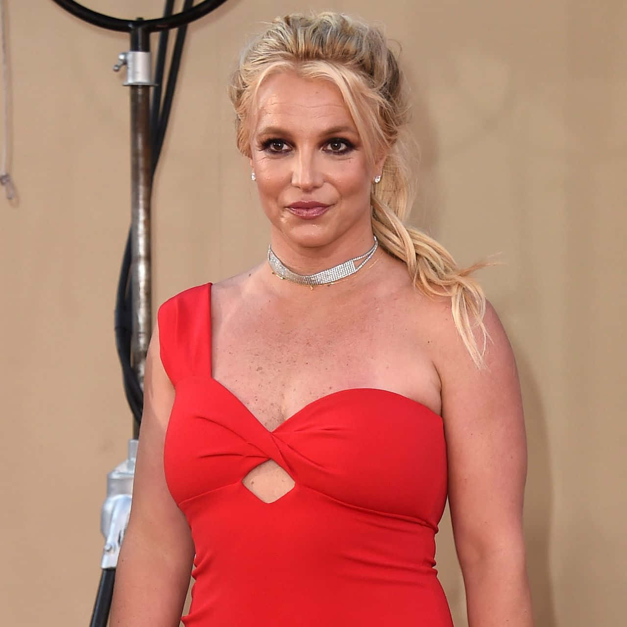 Britney Spears 桌布 壁紙 — Britney Spears 在米色背景前，身穿大膽的紅色單肩連衣裙，帶有鏤空細節和鑽石頸鍊，擺出姿勢。