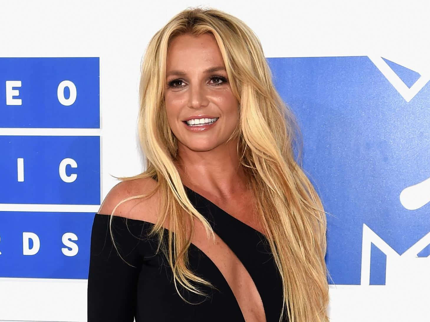 Britney Spears 桌布 壁紙 — Britney Spears 在藍色 MTV 音樂獎背景前，穿著黑色單肩裙燦爛微笑。
