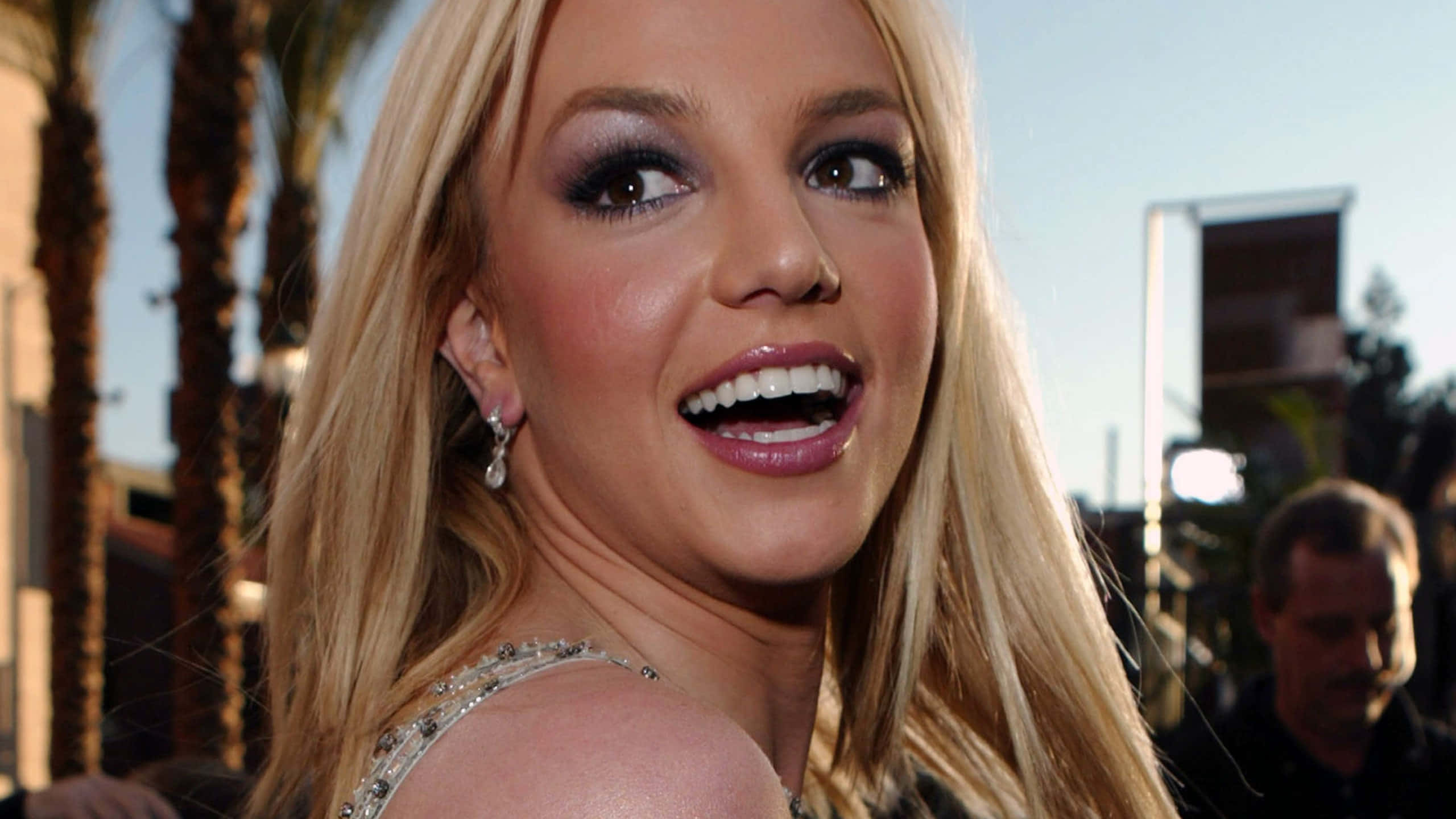 Britney Spears 桌布 壁紙 — Britney Spears 在戶外活動中，在柔和日光下，與棕櫚樹和建築物一起燦爛微笑。