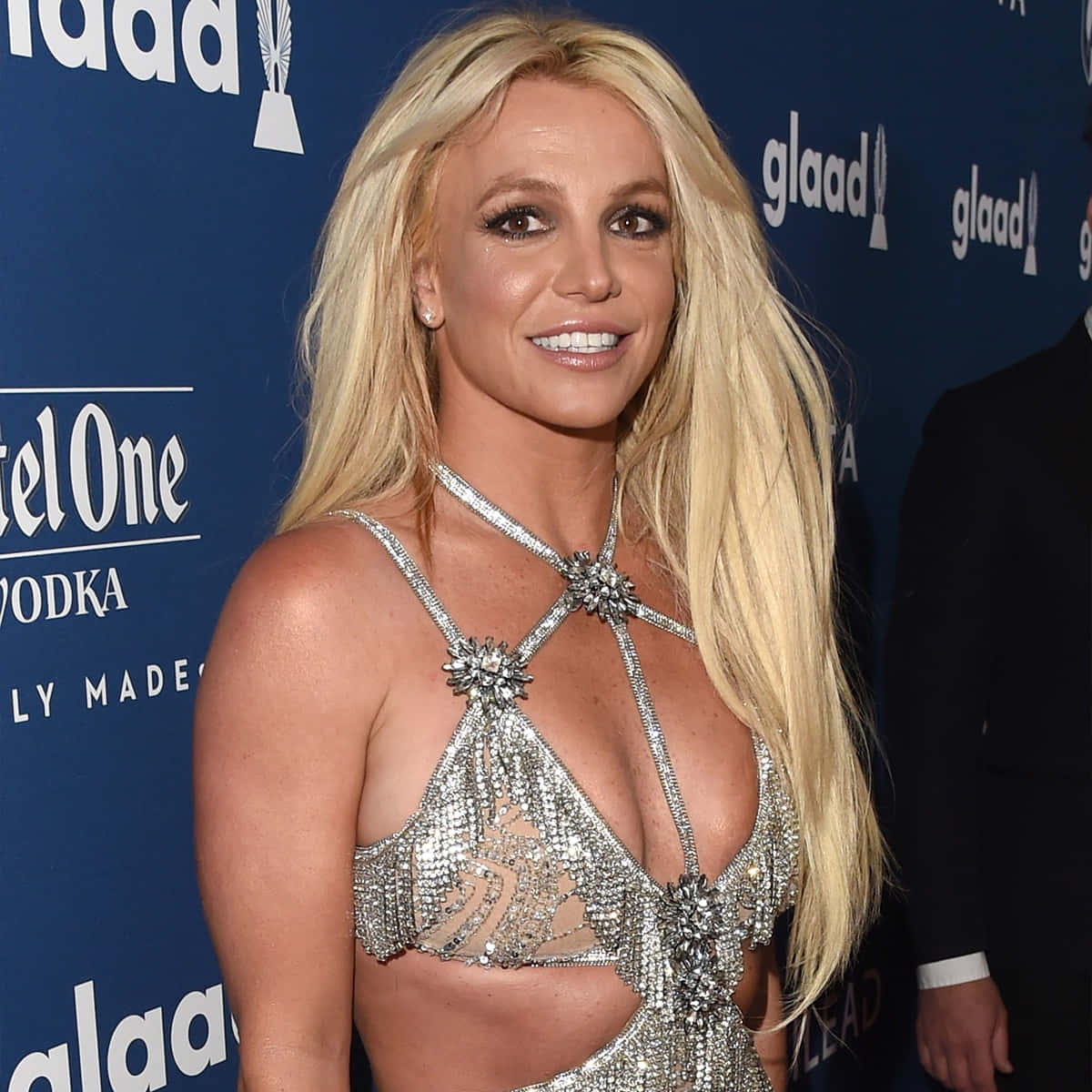 Britney Spears 桌布 壁紙 — Britney Spears 在GLAAD活動中穿著閃閃發光的銀色掛頸上衣，在藍色品牌背景前微笑。