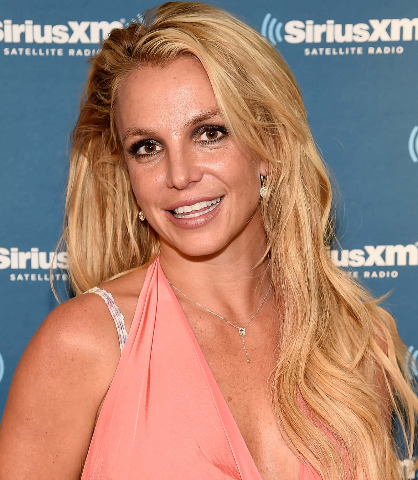 Britney Spears 桌布 壁紙 — Britney Spears 在藍色SiriusXM衛星廣播背景下，穿著珊瑚色掛頸上衣，露出燦爛笑容。
