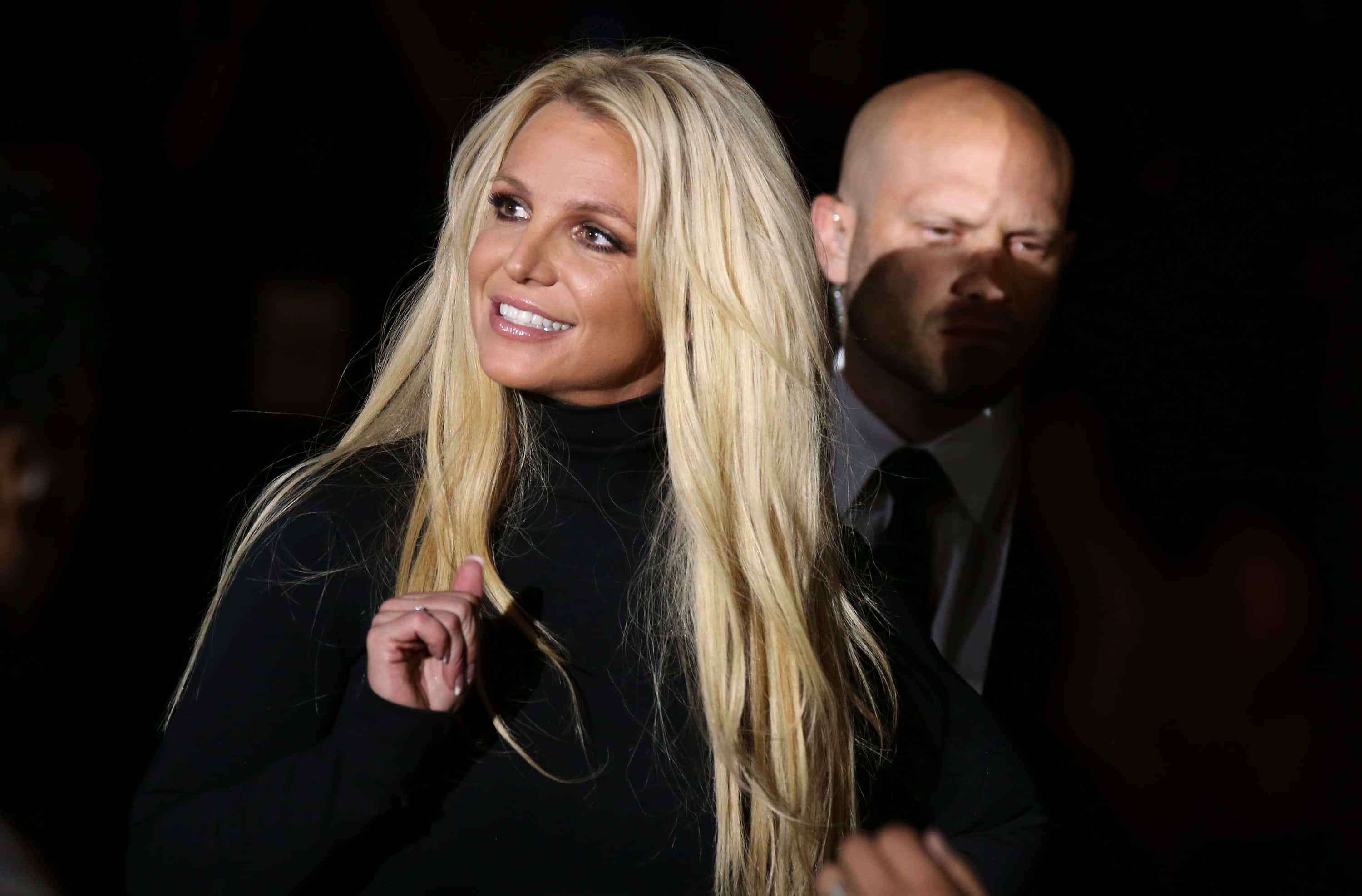 Britney Spears 桌布 壁紙 — Britney Spears 穿著黑色高領毛衣，在黑暗環境中燦爛微笑，身後站著一位穿西裝的光頭男子。