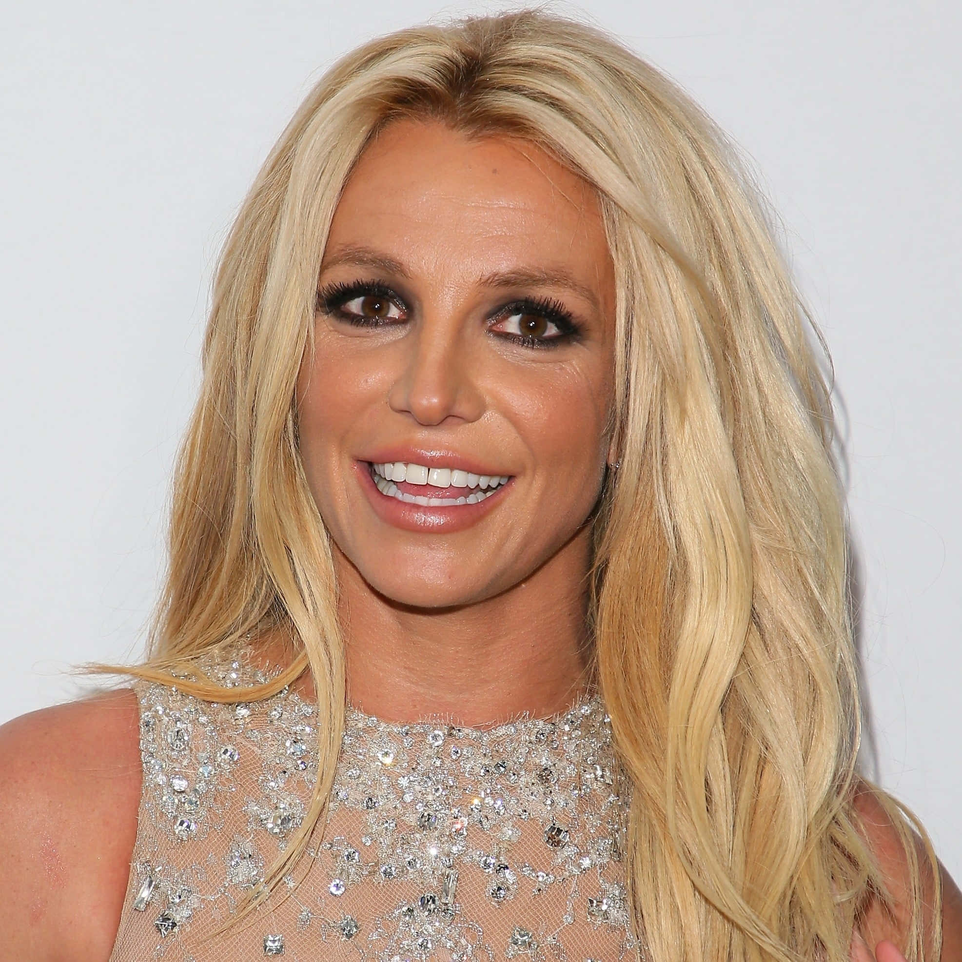 Britney Spears 桌布 壁紙 — Britney Spears 笑容燦爛，長長的金色頭髮，閃閃發光的裝飾上衣，背景是純白色。