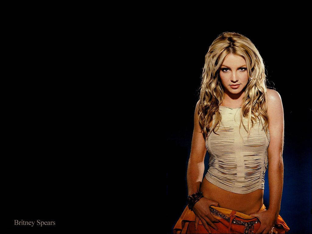 小甜甜布蘭妮 桌布 壁紙 — Britney Spears 自信地擺姿勢，身穿破爛的米色上衣和橙色裙子，背景為純黑色。