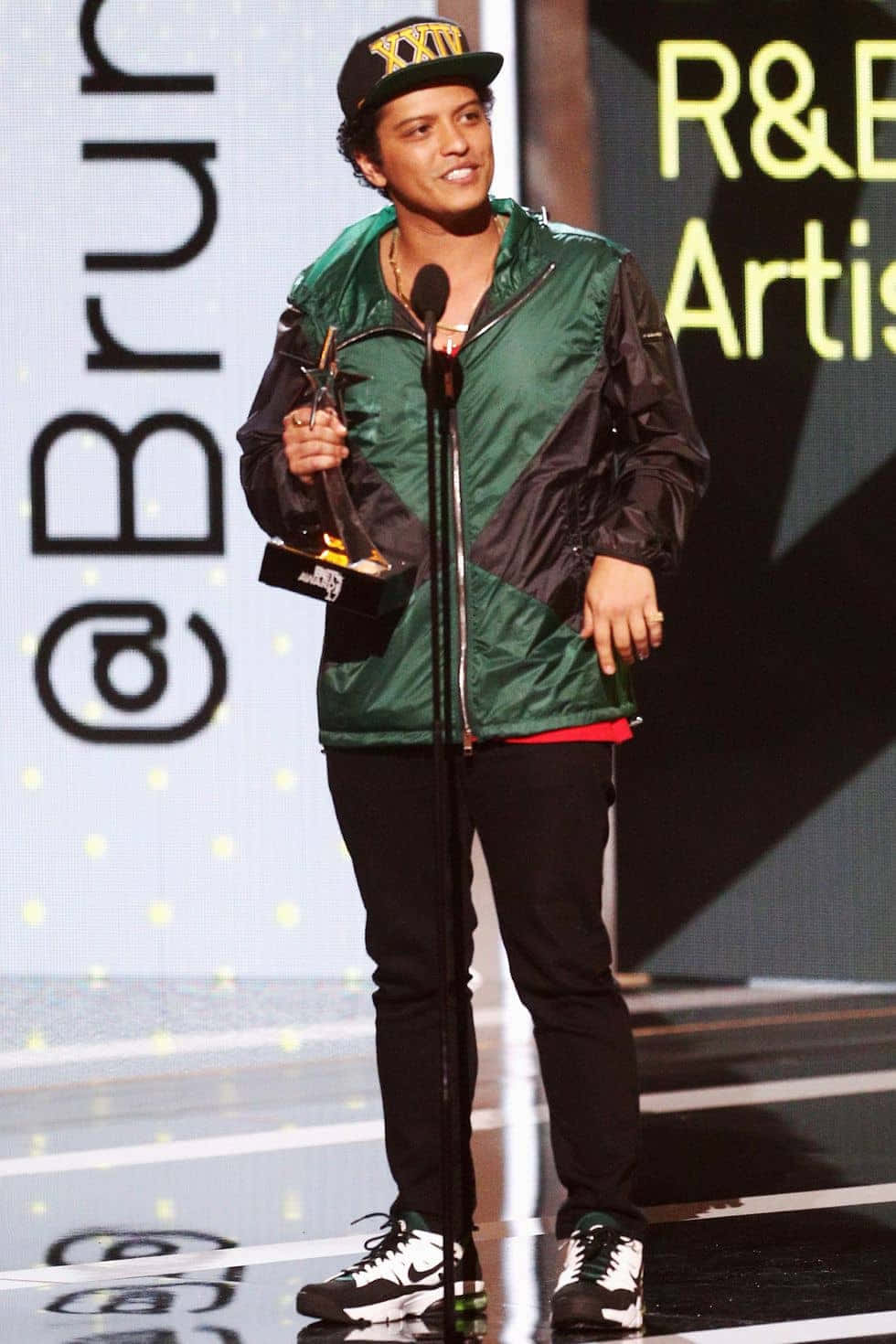 Bruno 桌布 壁紙 — Bruno Mars 站在舞台上，手持獎項，身穿綠色和黑色外套及帽子，在明亮的舞台燈光下。