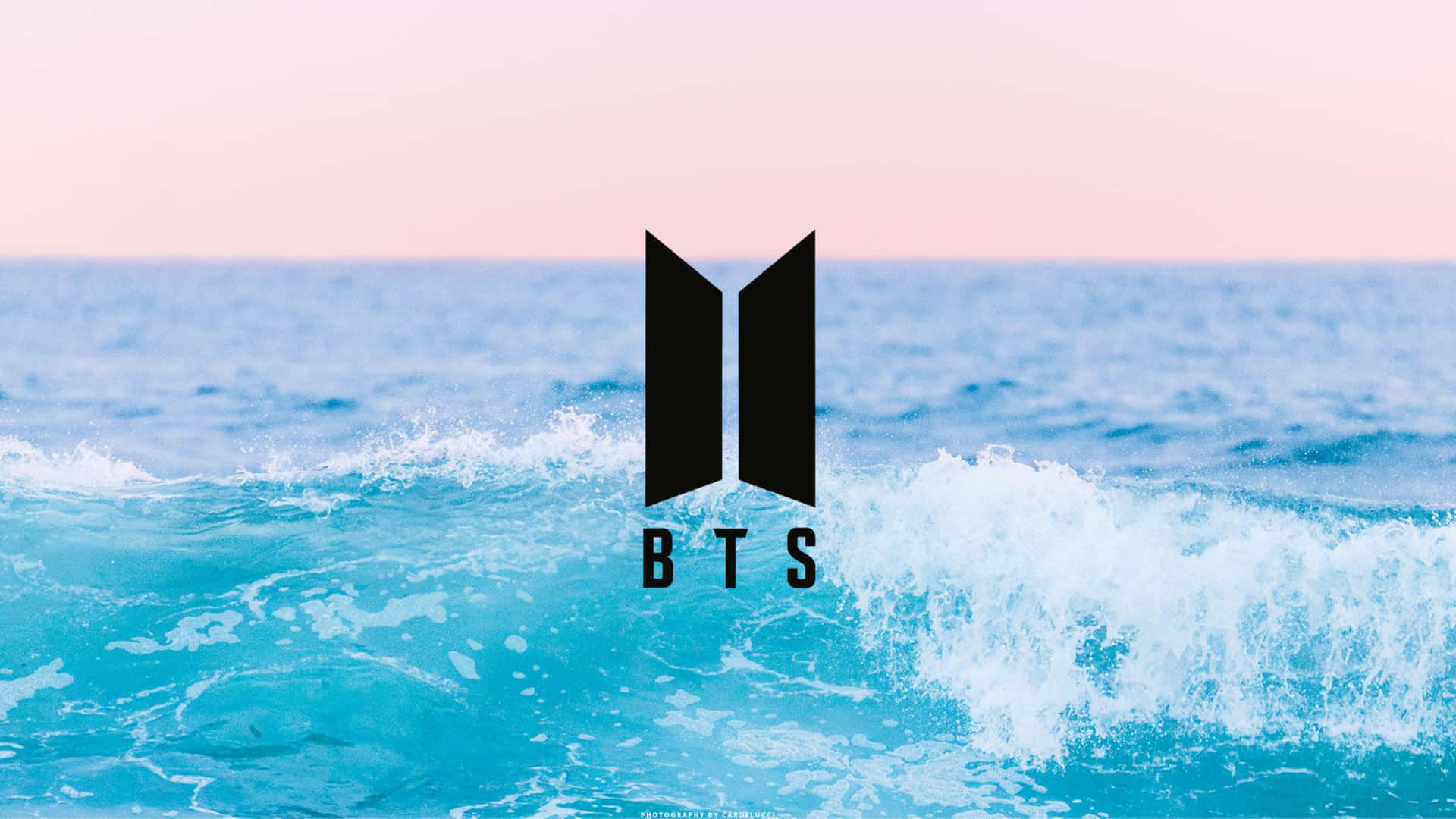 Pastel Blue Aesthetic Desktop 桌布 壁紙 — BTS 標誌在柔和粉紅色的天空下，大膽地矗立在充滿活力的綠松色海浪之上。