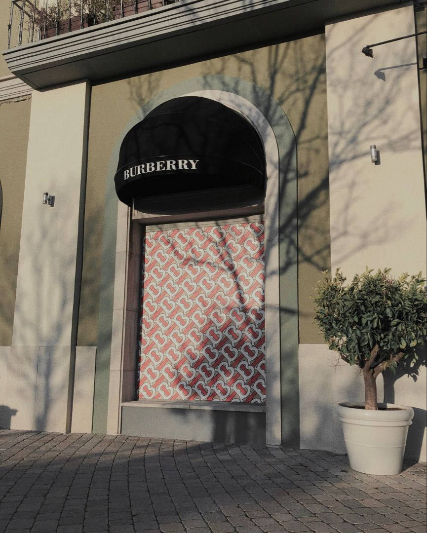 Burberry 桌布 壁紙 — Burberry 店鋪外牆，設有黑色遮陽篷及圖案櫥窗展示，在石行人道上投射樹影。