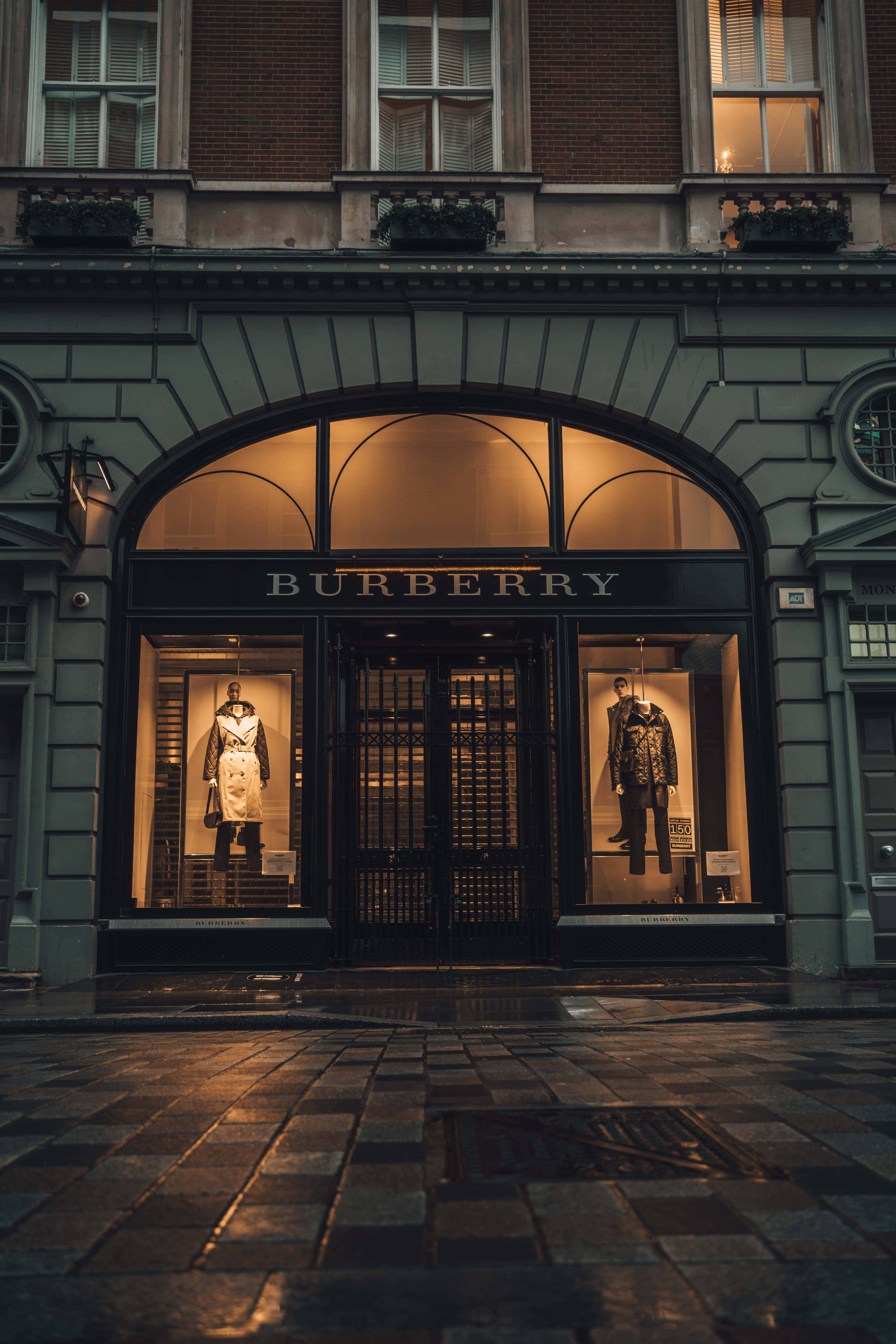 Burberry 桌布 壁紙 — Burberry 店鋪正面在黃昏時溫暖地發光，拱形窗戶中展示穿著風衣的人體模型，背景是濕漉漉的鵝卵石街道。