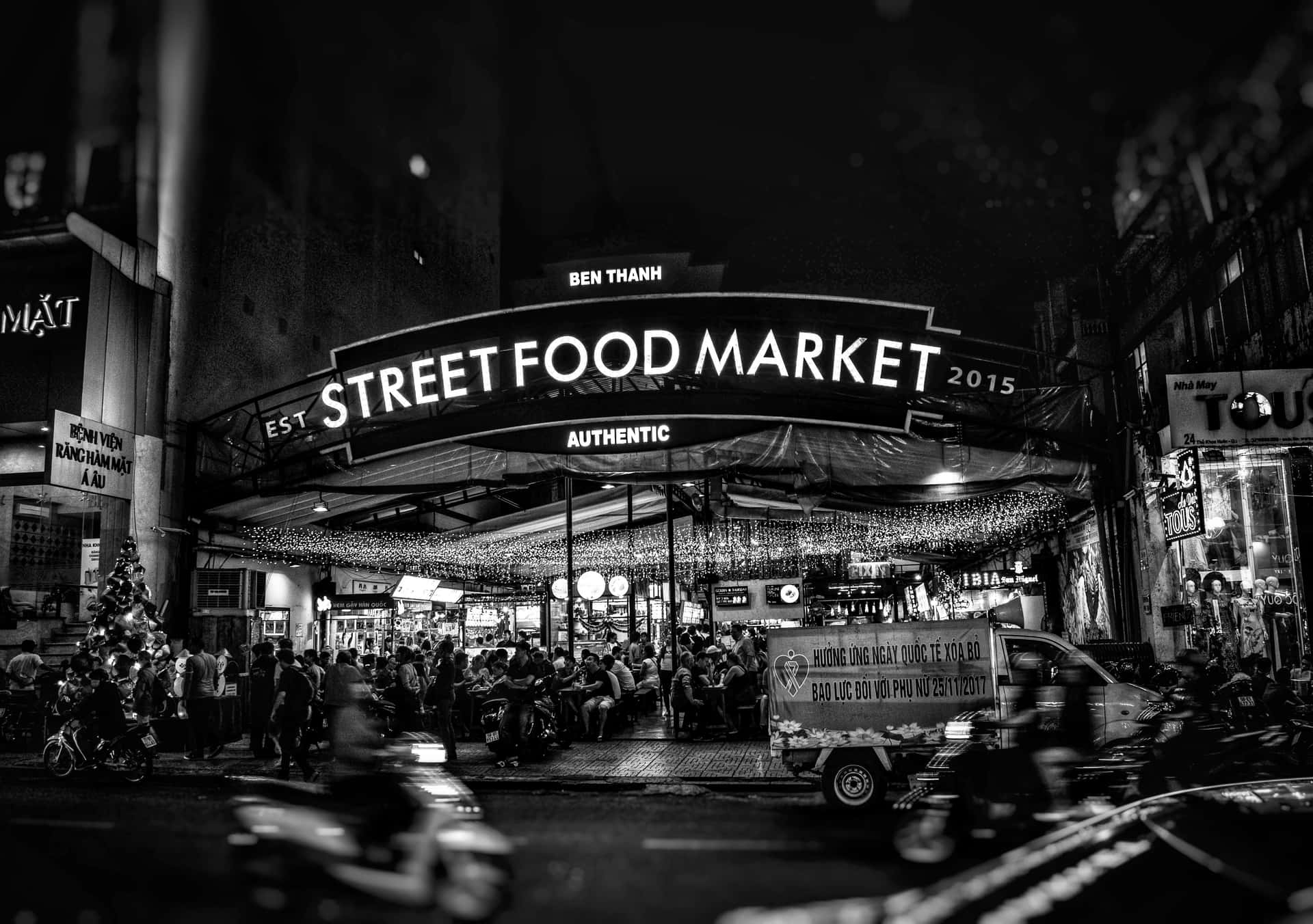 壁紙 — 濱城街頭美食市場熱鬧的黑白夜景，人群在發光招牌下用餐，機車穿梭而過。