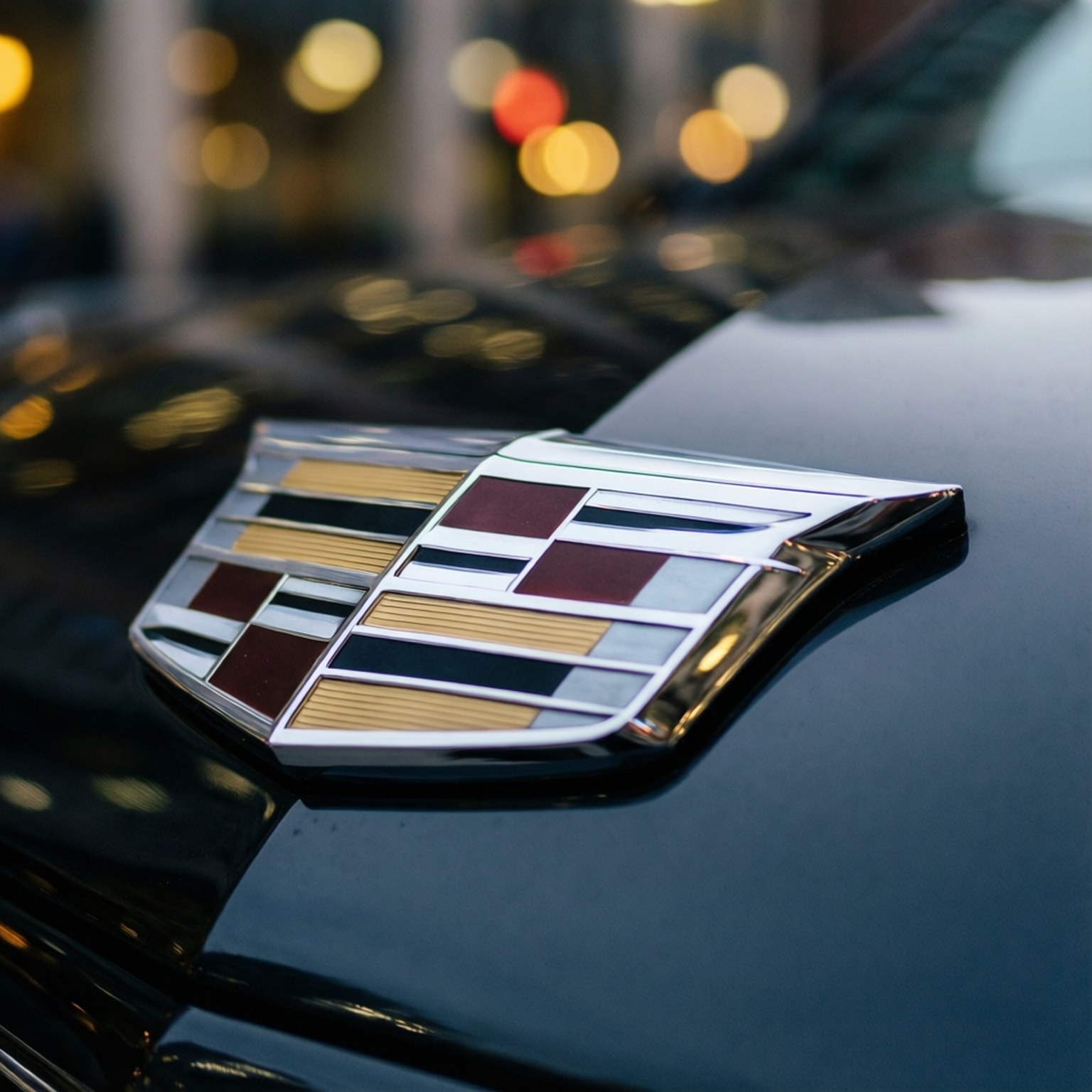 Emblem of Luxury 壁紙 — 凱迪拉克徽章在黑色引擎蓋上閃耀的特寫，背景是模糊的城市燈光。