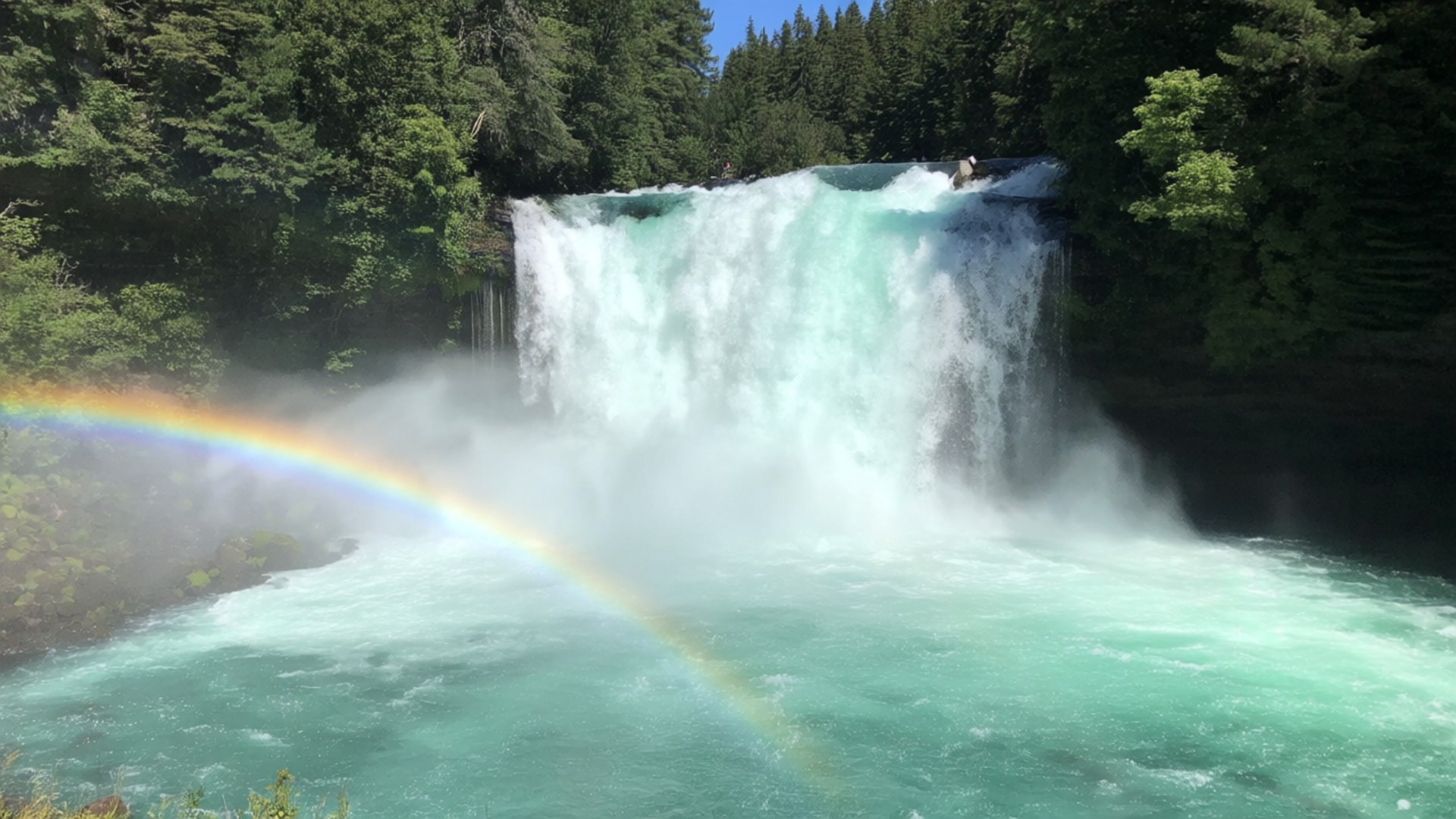 Cascading Waterfall 壁紙 — 一道鮮豔的彩虹拱在咆哮的綠松石瀑布之上，周圍是茂密的綠色森林，天空清澈湛藍。