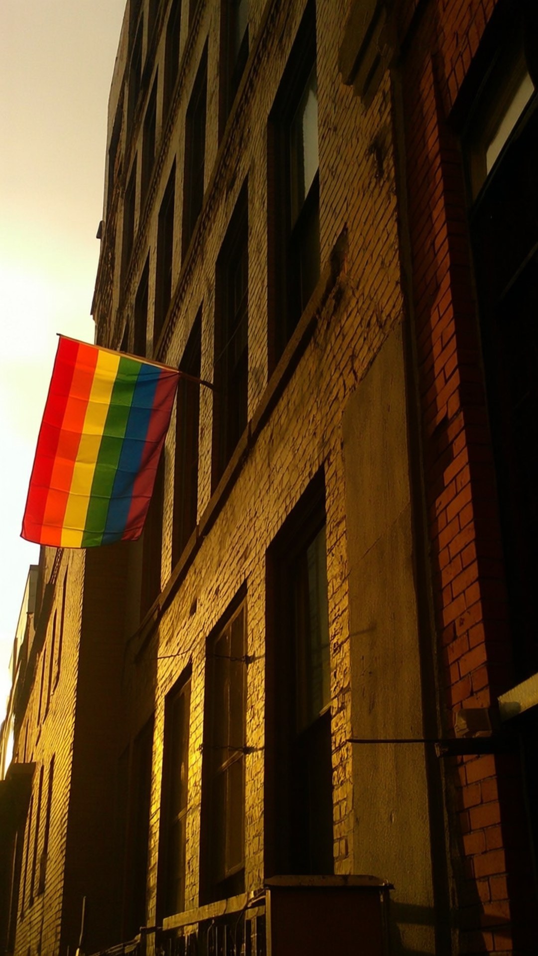 Urban Pride Window 壁紙 — 一面彩虹旗在黃金時段的陽光照射下，在磚砌建築外牆上飄揚，投下長長的影子。
